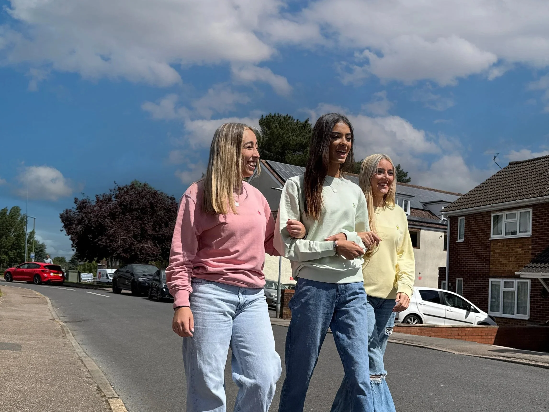 Three young women walking arm-in-arm on a residential street, smiling and enjoying a sunny day with a partly cloudy sky.