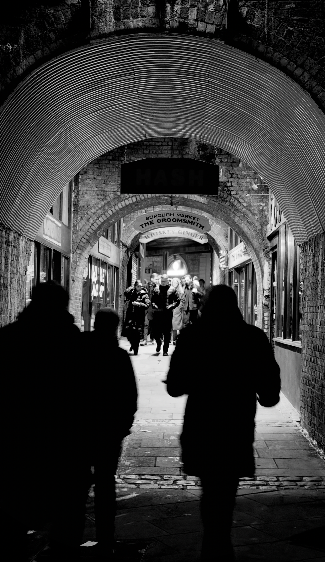 People walking through a covered alleyway with brick walls and arched ceilings, storefronts on each side, and signs hanging overhead, including one that reads 'Borough Market The Groomsmit' and another with partially visible text.