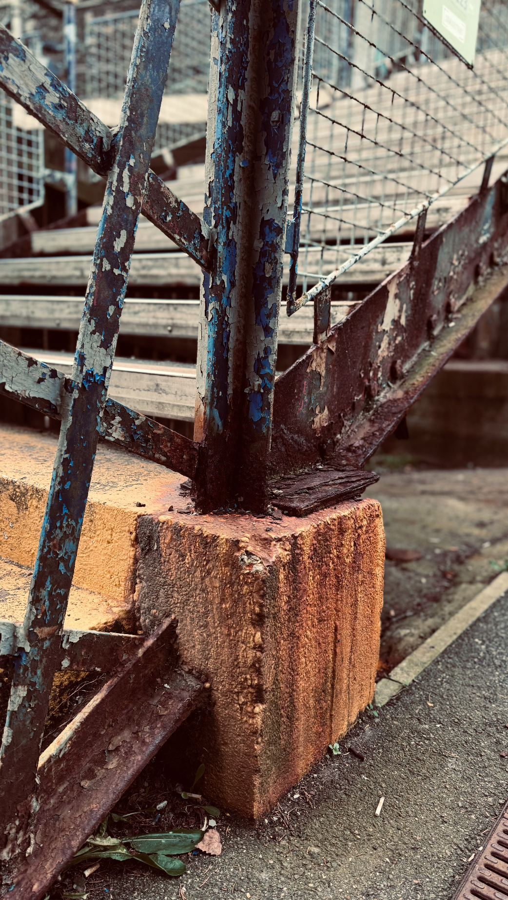 Close-up of a rusty metal staircase with peeling blue paint, attached to a weathered concrete block, next to an asphalt pavement.