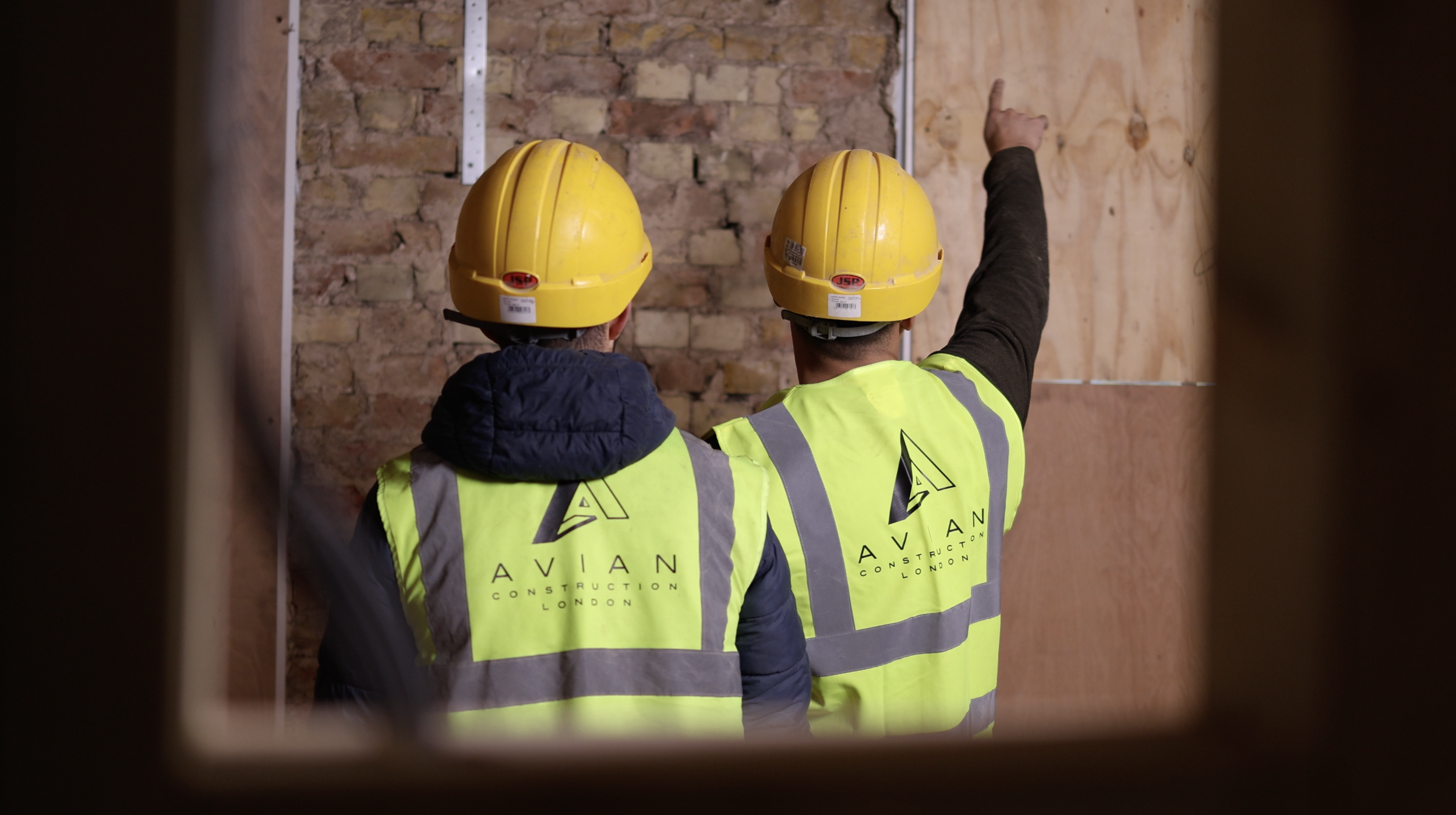 Two construction workers wearing yellow hard hats and high-visibility vests, one pointing at a wall in an indoor construction site.