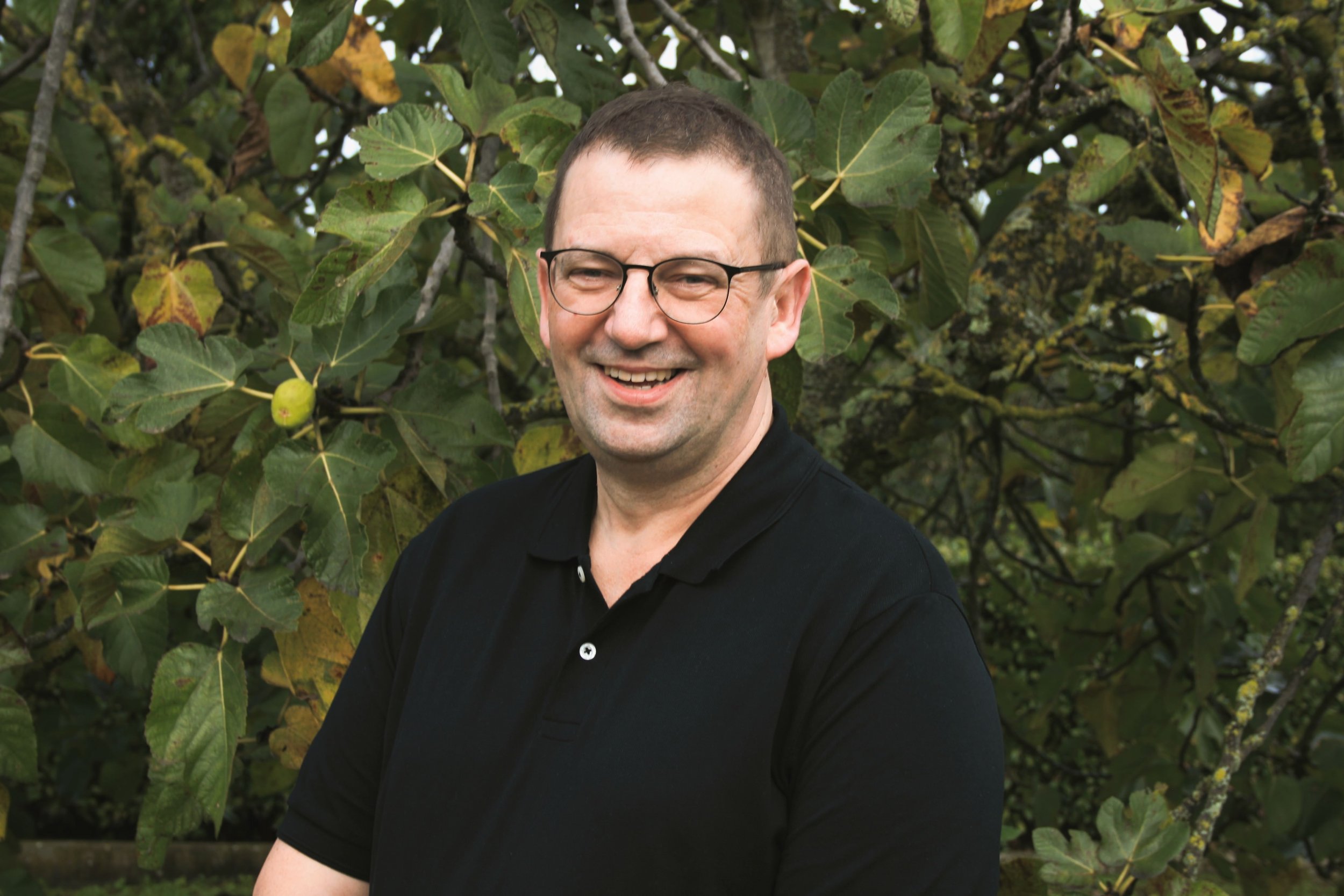 A man with glasses smiling outdoors in front of a leafy tree, wearing a black polo shirt.
