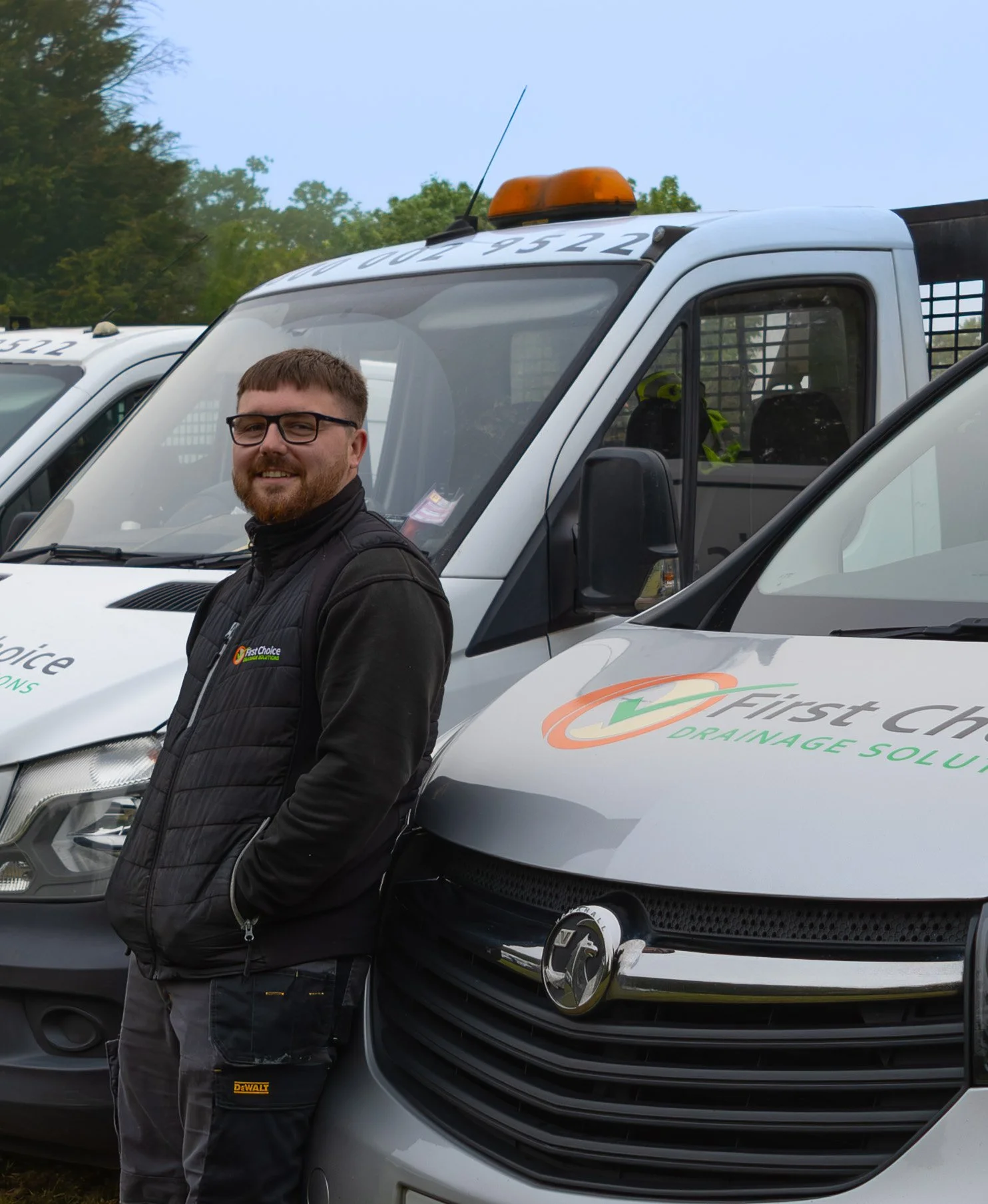 A man with glasses and a beard standing in front of a white utility truck with the logo 'First Choice Drainage Solutions' visible on the side. The man is wearing a black vest and gray pants with a DeWalt tool pocket. There are more similar vehicles in the background with trees and a cloudy sky.