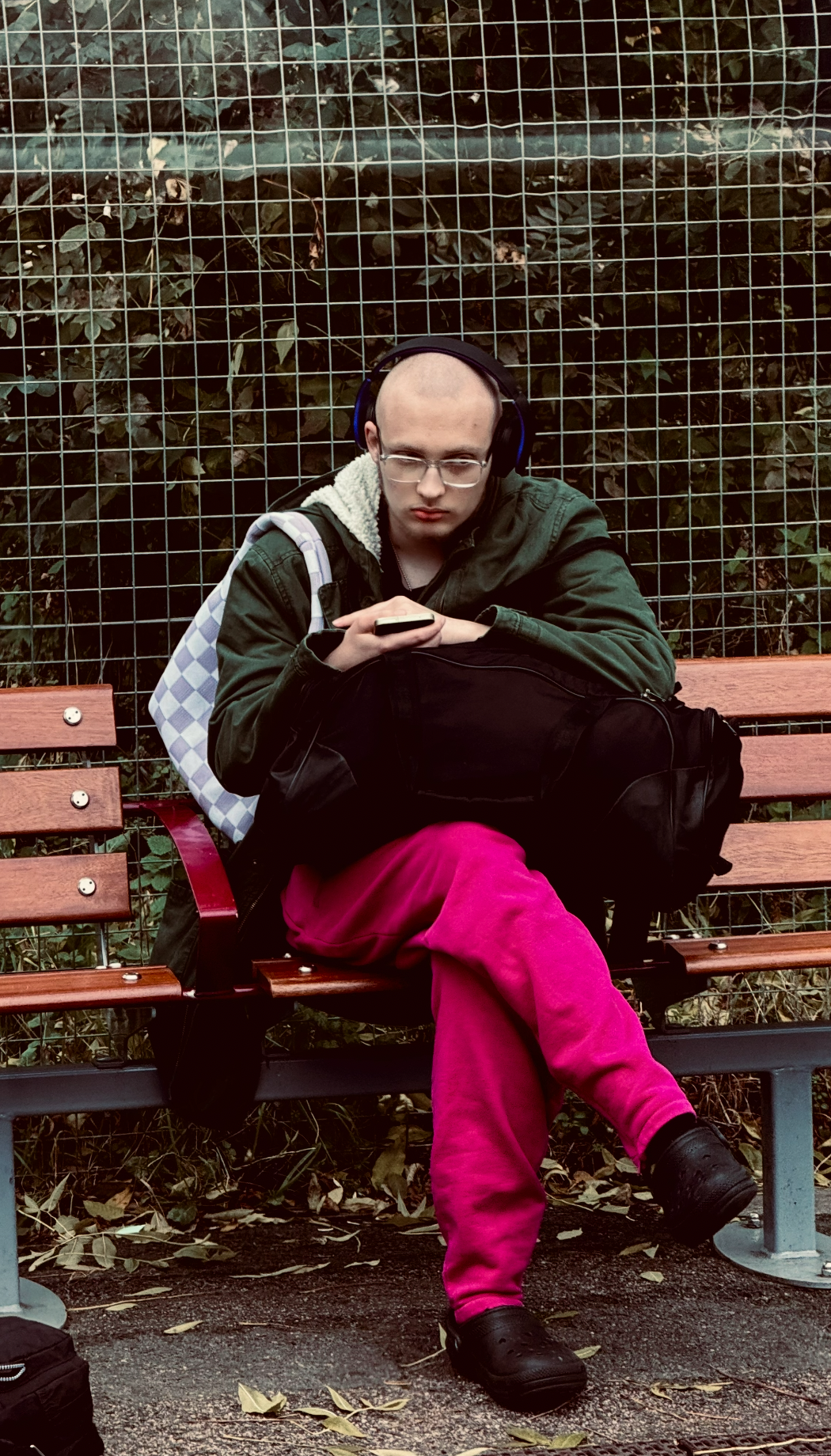 Young man with glasses wearing headphones, green jacket, and pink pants sitting on a park bench looking at his phone, with a backpack beside him and a wire mesh fence and foliage in the background.