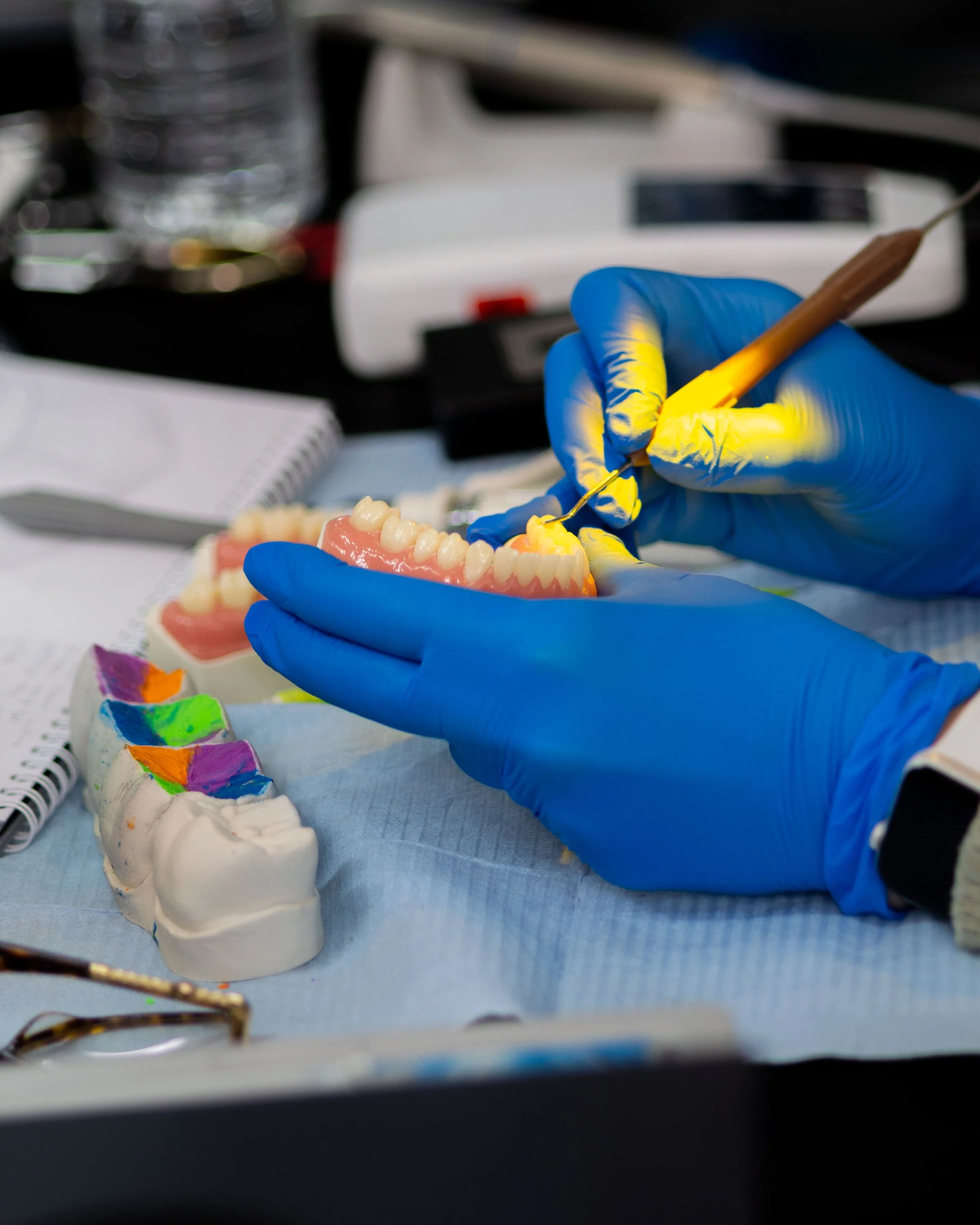 A person wearing blue gloves holding a dental model and a dental instrument, with dental tools and a water bottle in the background.