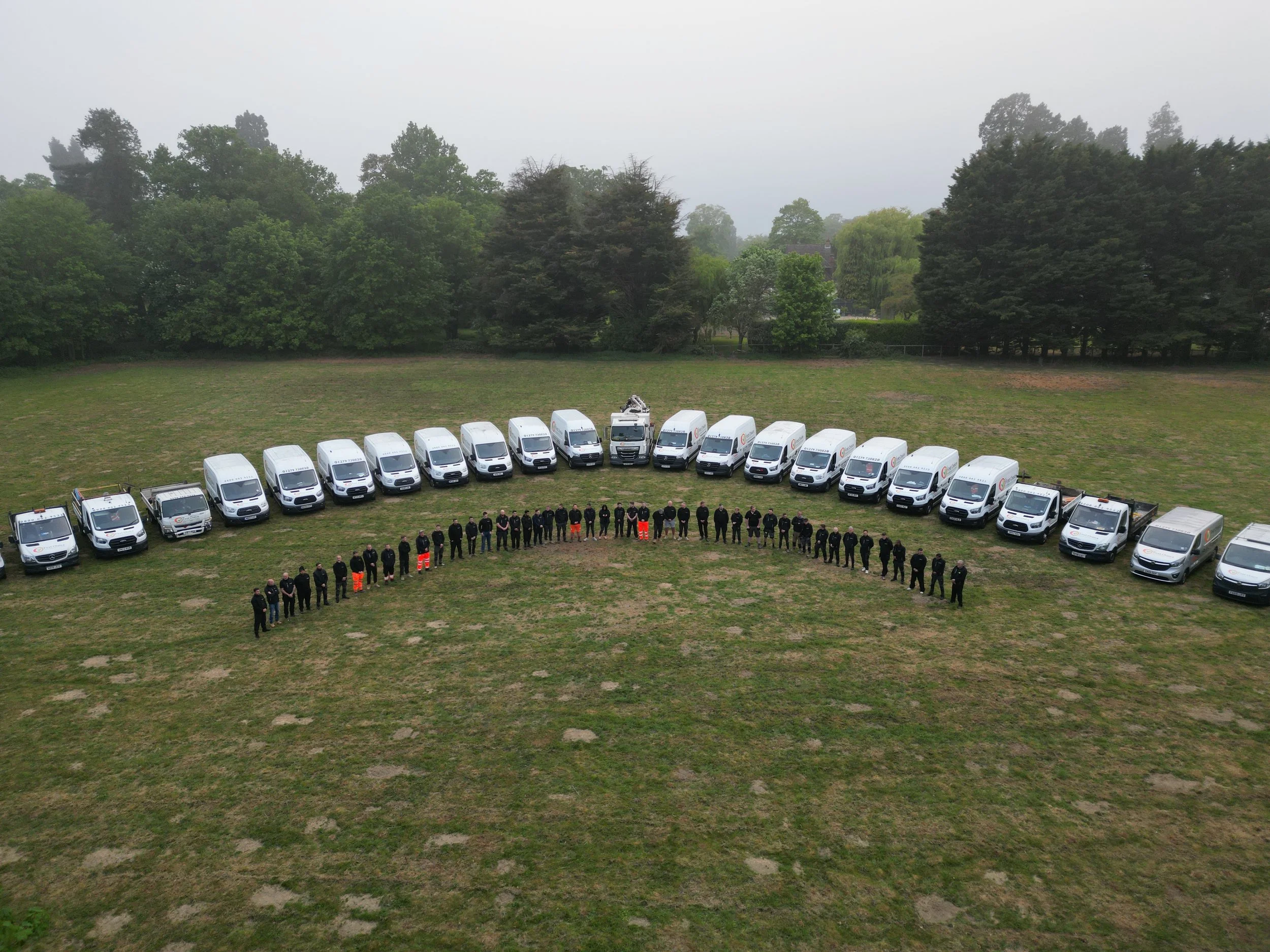 A group of uniformed workers standing in a line in front of a row of white work vans on a grassy field with trees in the background.