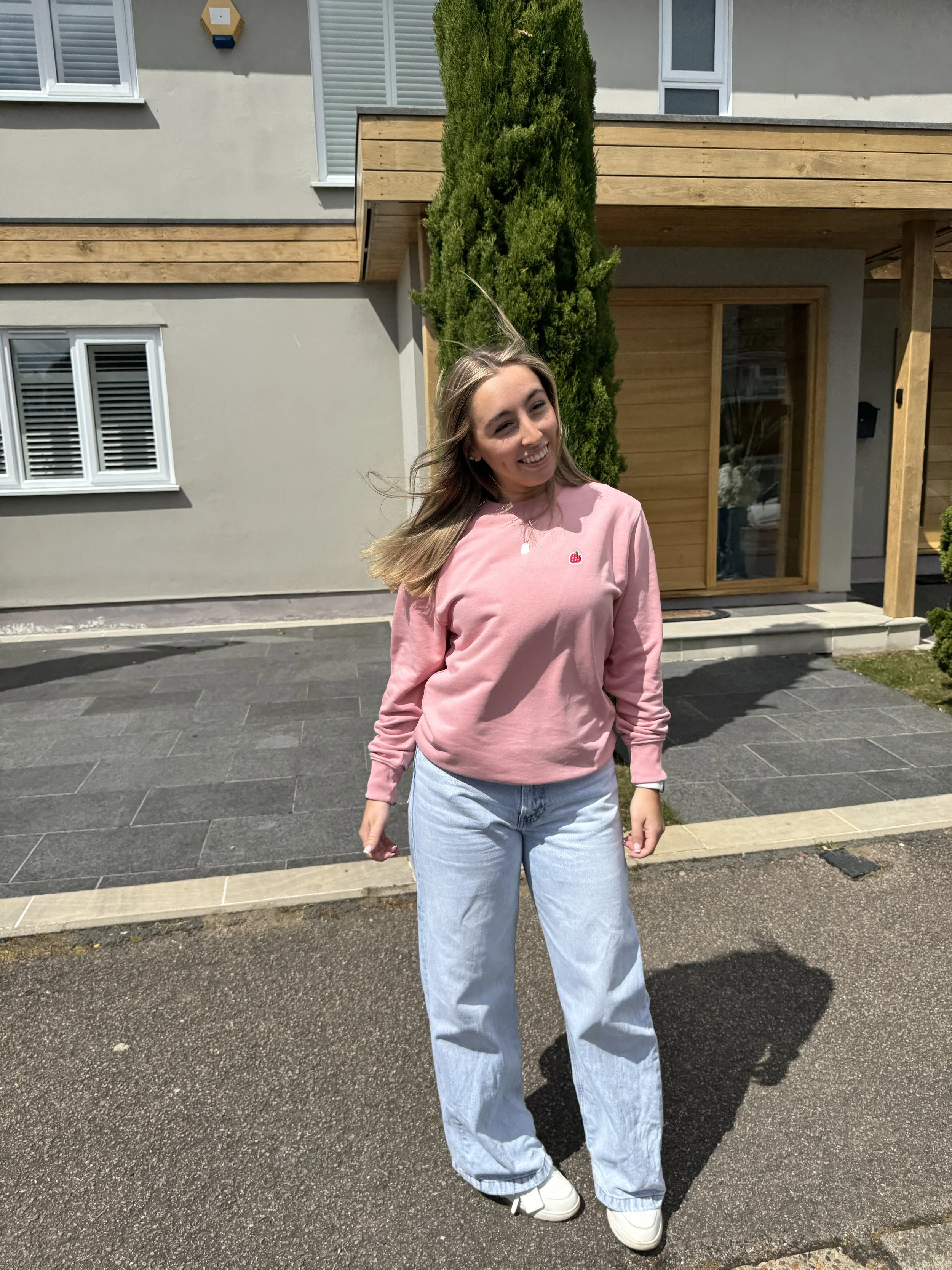A young woman with light brown hair, wearing a pink sweatshirt with a small red embroidered strawberry, light blue jeans, and white sneakers, smiling outdoors on a sunny day in front of a modern house with a wooden and gray exterior and a green tree.