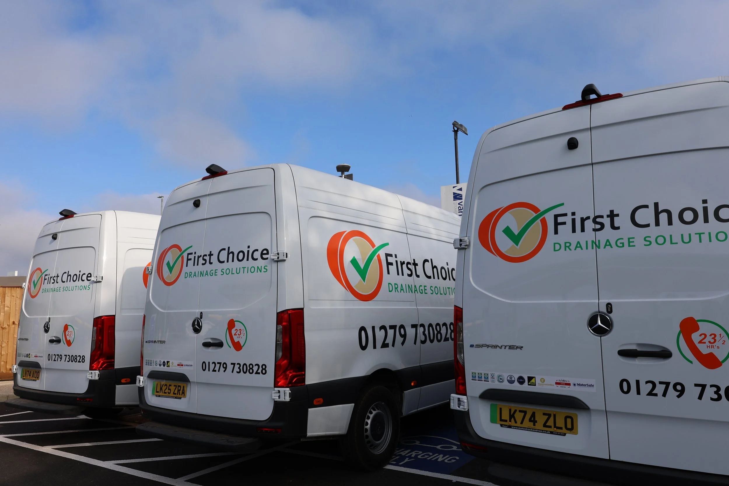 Three white delivery vans parked in a lot, bearing the logo and contact information for First Choice Drainage Solutions. The vans are positioned side by side, with clear signage and license plates visible.