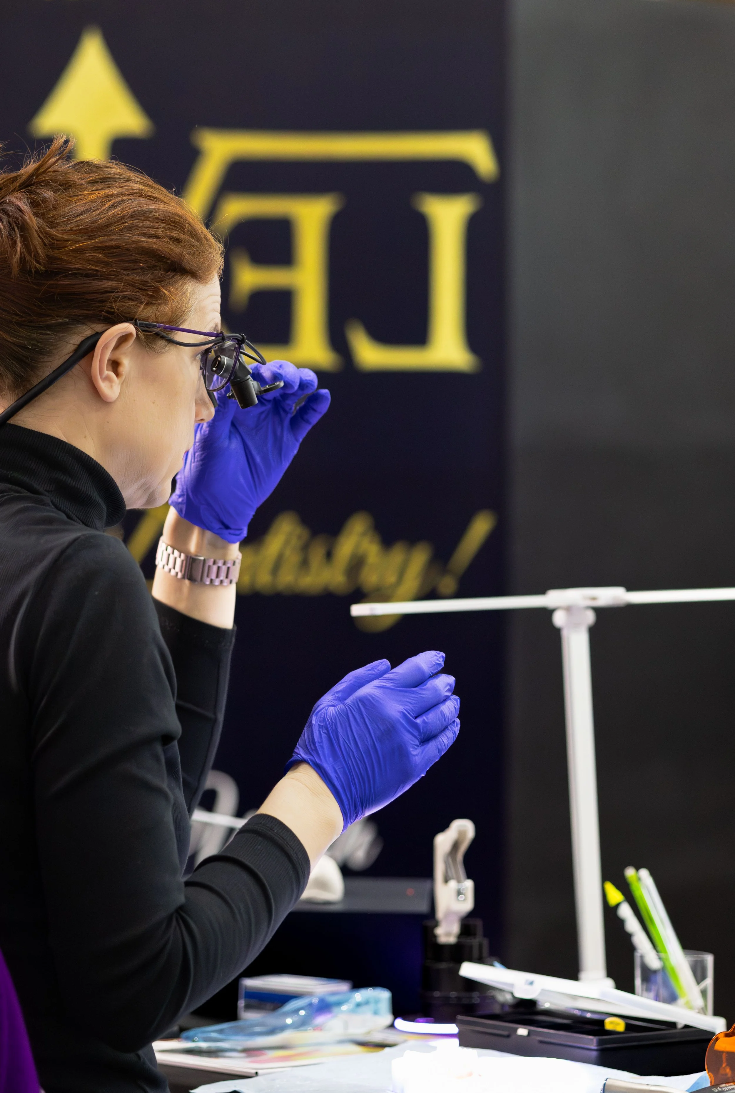 A scientist wearing purple gloves and safety glasses looks through a magnifying device at a laboratory table with scientific equipment, with a background featuring a dark blue wall and yellow lettering.