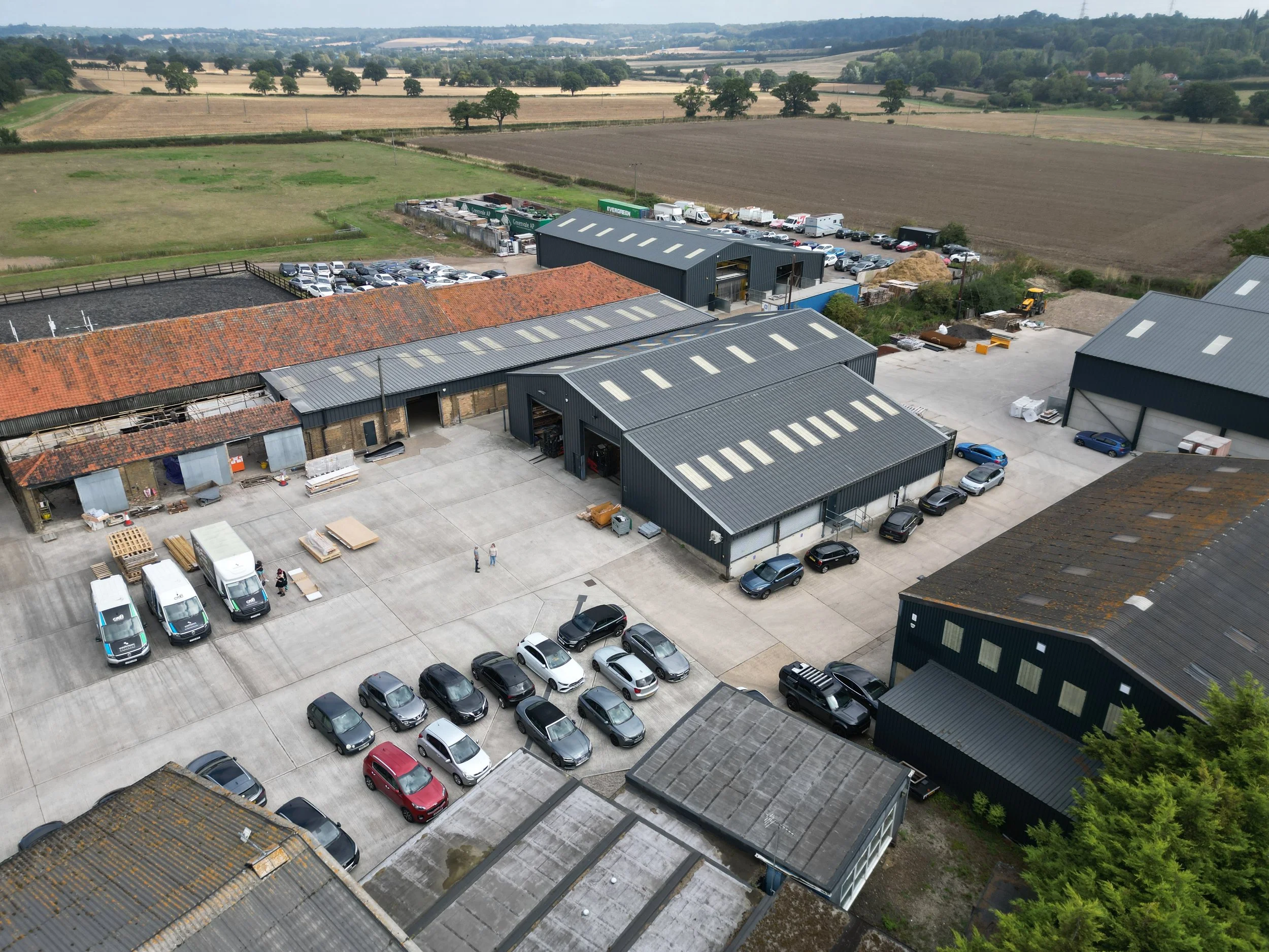 Aerial view of a complex of industrial and warehouse buildings with cars parked outside, surrounded by open fields and farmland.