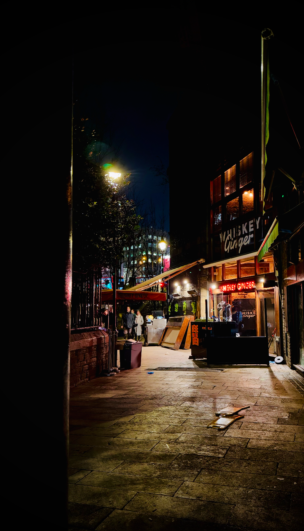 Nighttime street scene outside Whiskey Ginger bar with bright neon signs, outdoor tables, and two people walking.