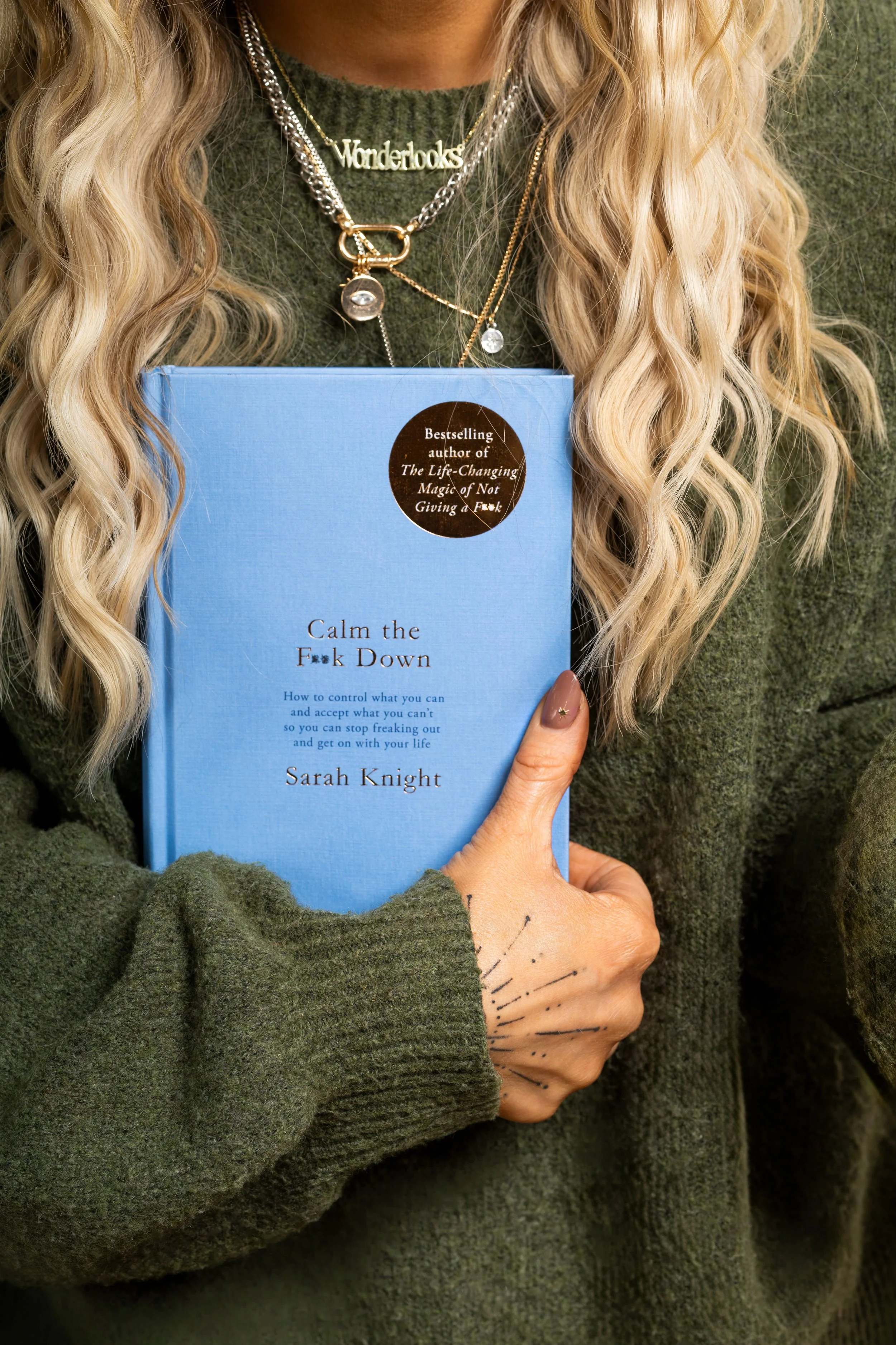 A woman with long, wavy blonde hair is wearing multiple necklaces, including one with the word 'Wonderlooks,' and is holding a blue book titled 'Calm the F*ck Down' by Sarah Knight. She is dressed in an olive green sweater.