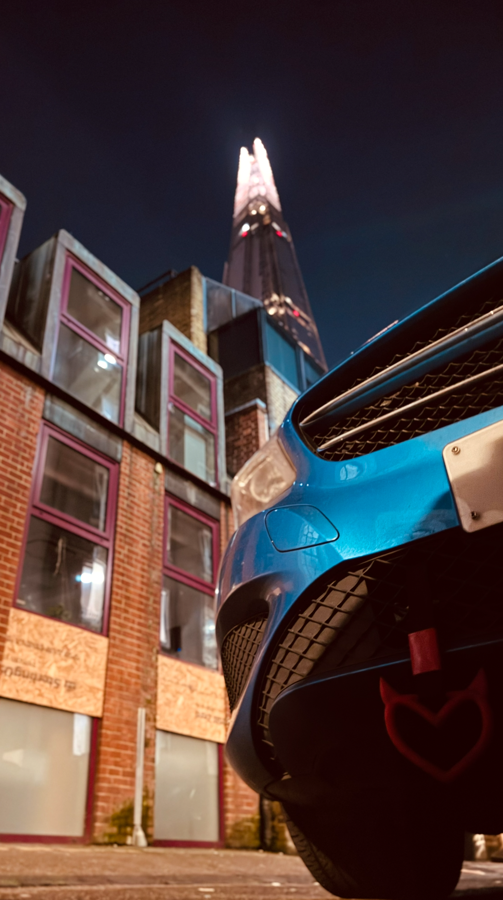 Low-angle night photo of a blue sports car in front of modern brick and glass buildings, with a tall illuminated skyscraper in the background.