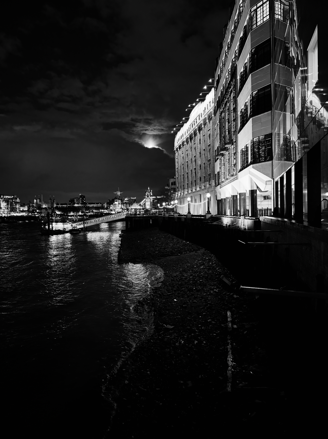 Nighttime black and white photo of a city waterfront with modern buildings, bridge, and distant illuminated skyline under cloudy sky.