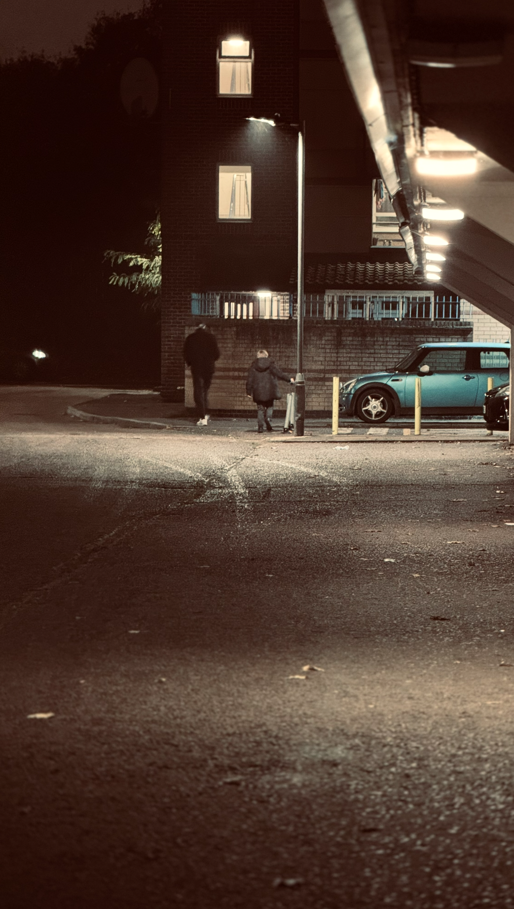 Nighttime scene of two people walking in a parking lot near a brick building, with a blue Mini Cooper parked on the right and bright overhead lights illuminating the area.