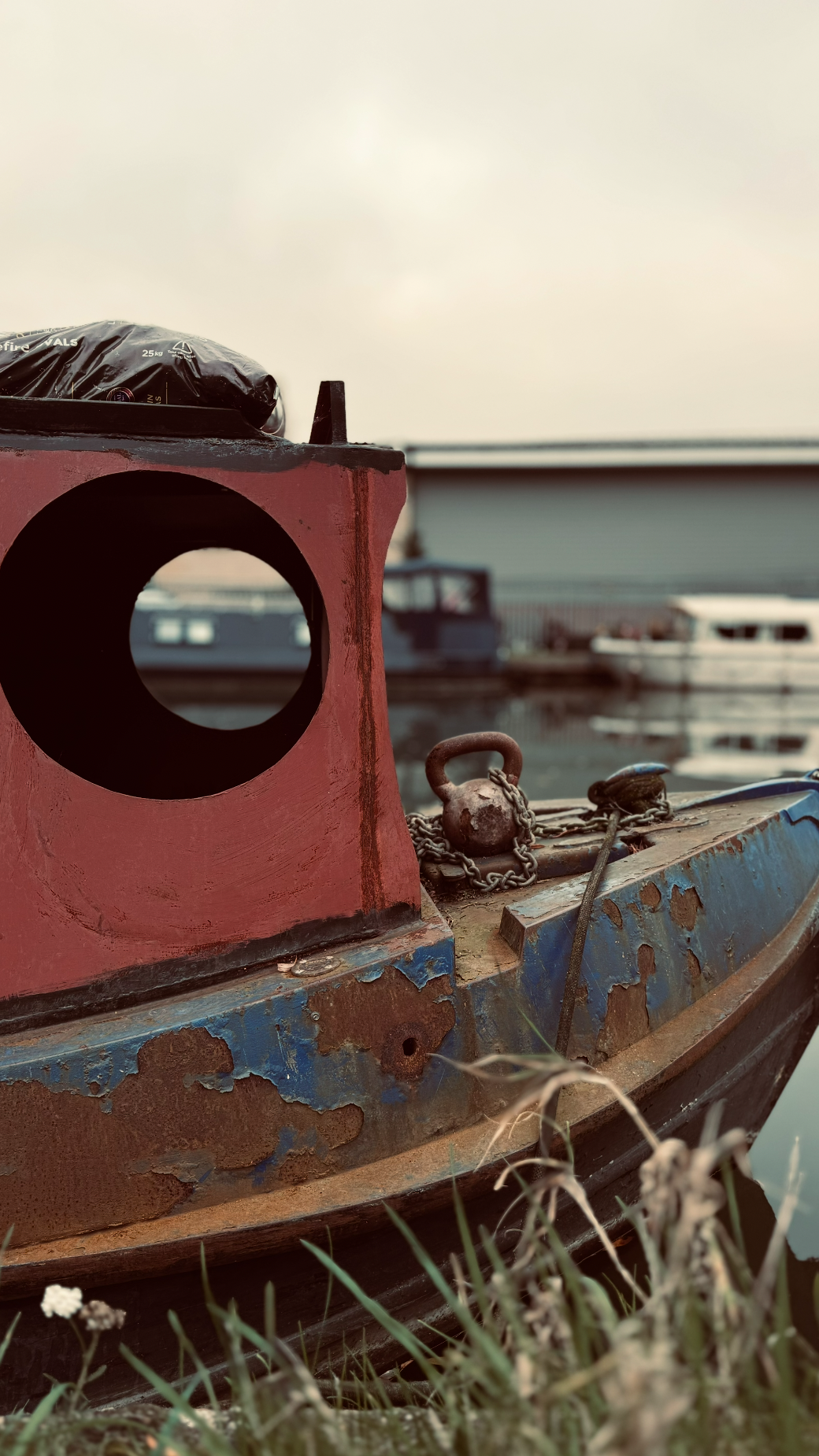 Close-up of a weathered, rusty boat with chain and hook, docked near a marina with boats in the background.