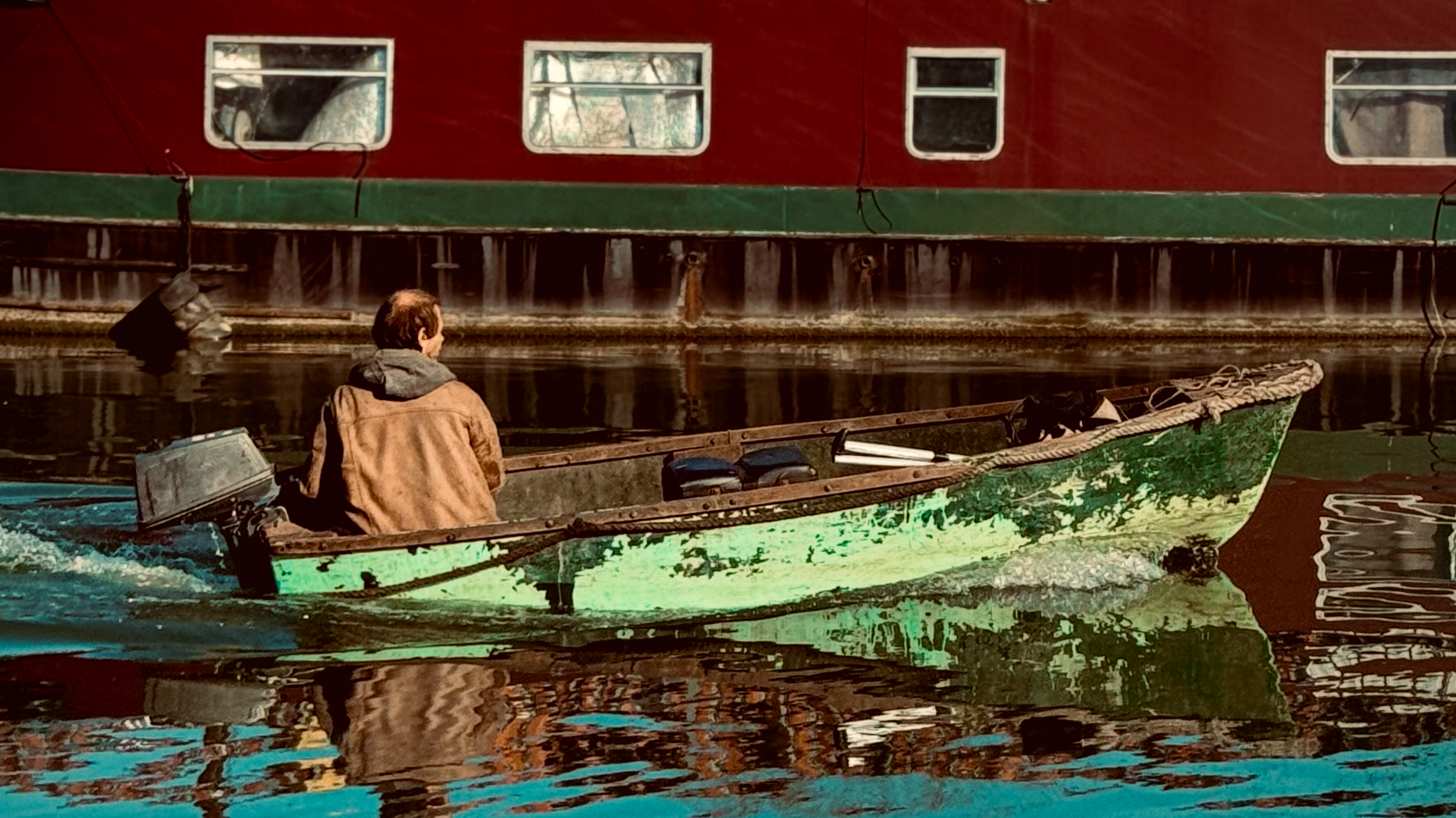 A person in a brown jacket and gray hoodie sitting in a small boat on water near a large red boat with multiple windows.