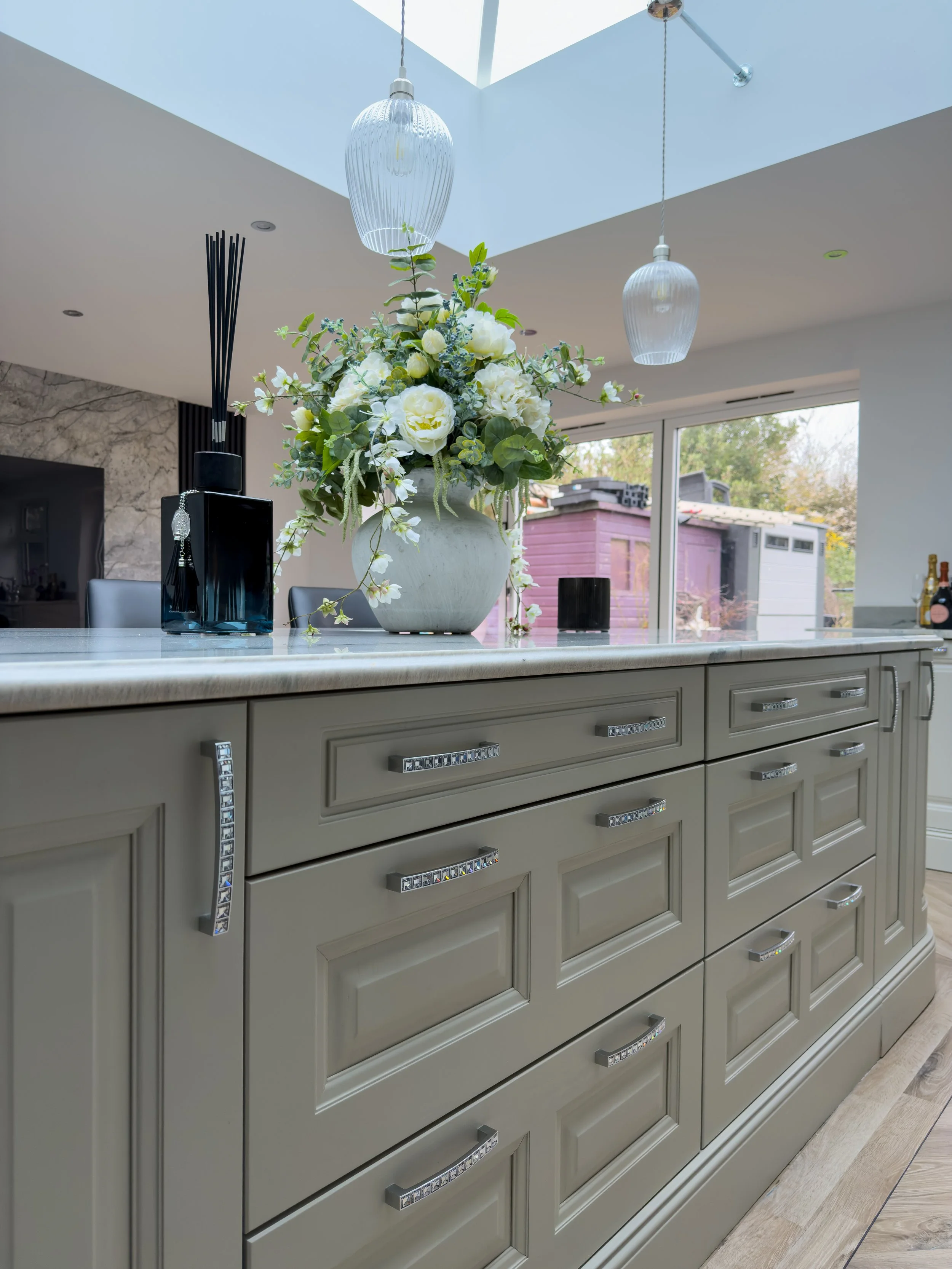 A kitchen island with beige drawers featuring decorative handles, topped with a large floral arrangement in a white vase, and two hanging pendant lights. A sliding glass door and outdoor scenery are visible in the background.