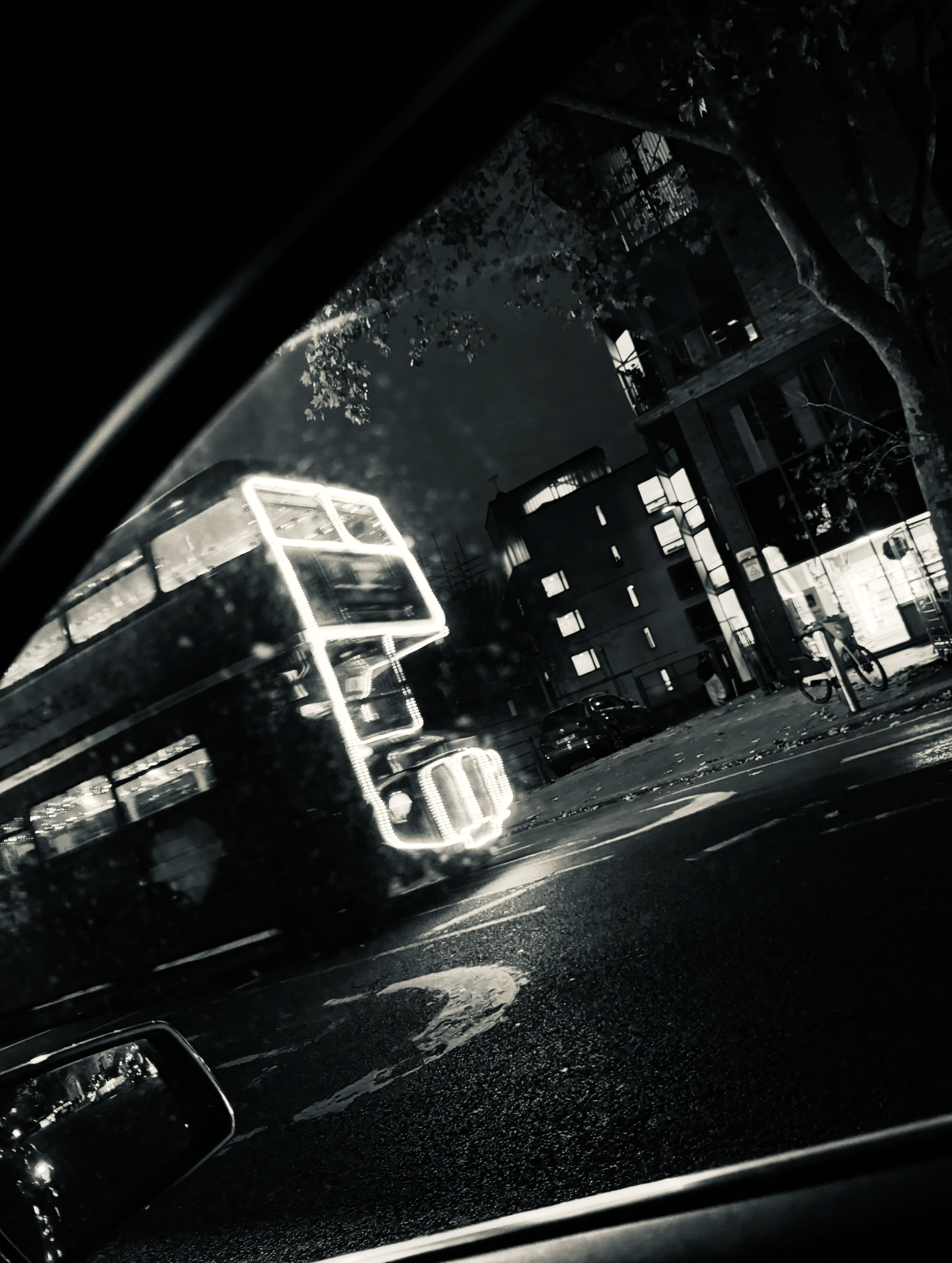 Nighttime view of a city street with illuminated neon sign shaped like a bus, seen through a car window with raindrops on the glass.