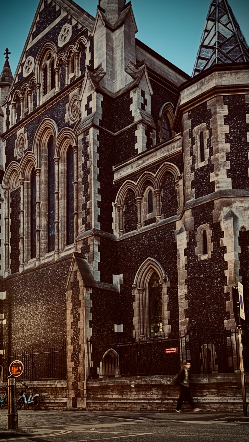 A historic Gothic-style church with intricate stonework and tall arched windows, located on a city street with a person walking past and bicycles parked nearby.