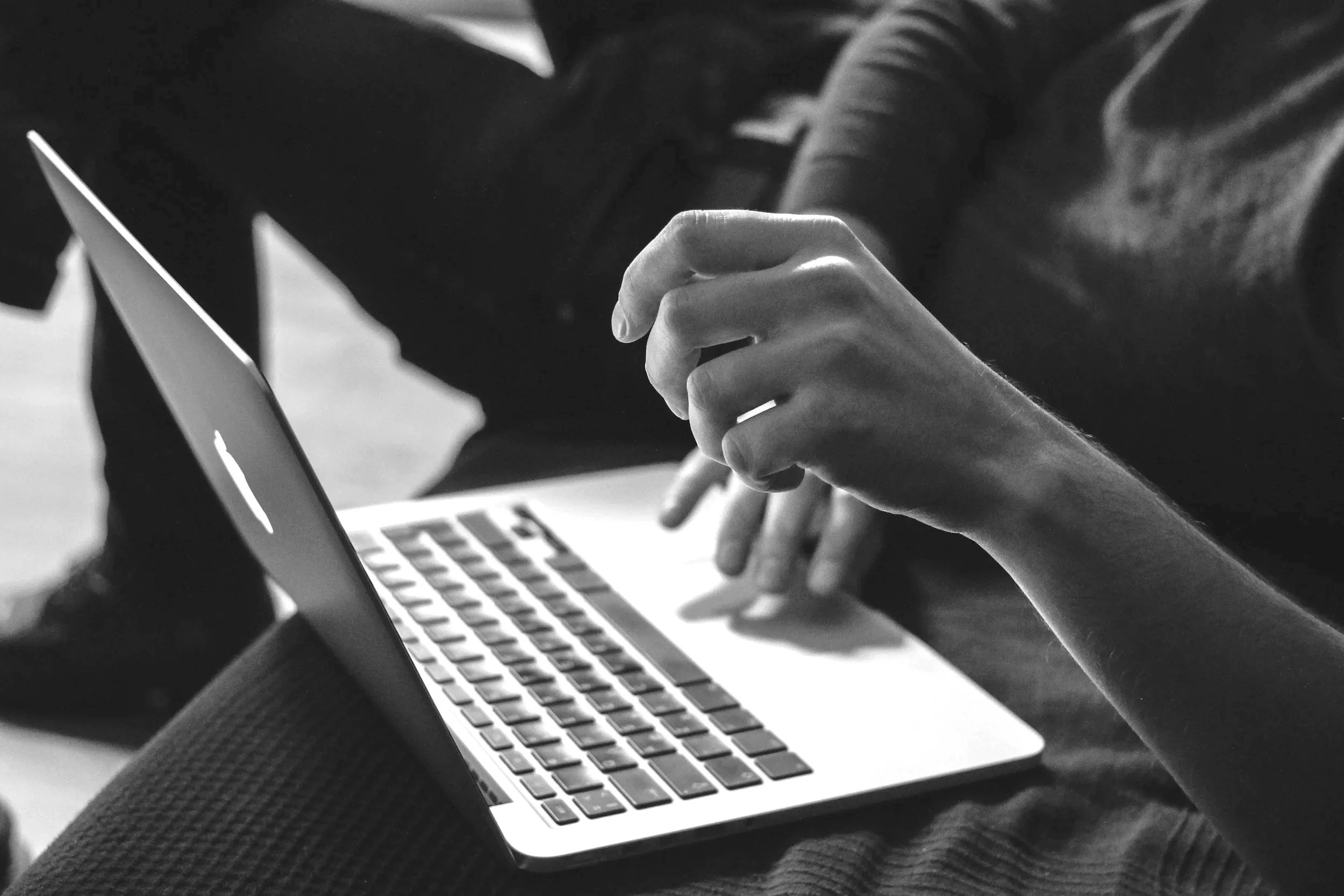 Close-up of a person's hand interacting with a laptop, with other people in the background, in black and white.