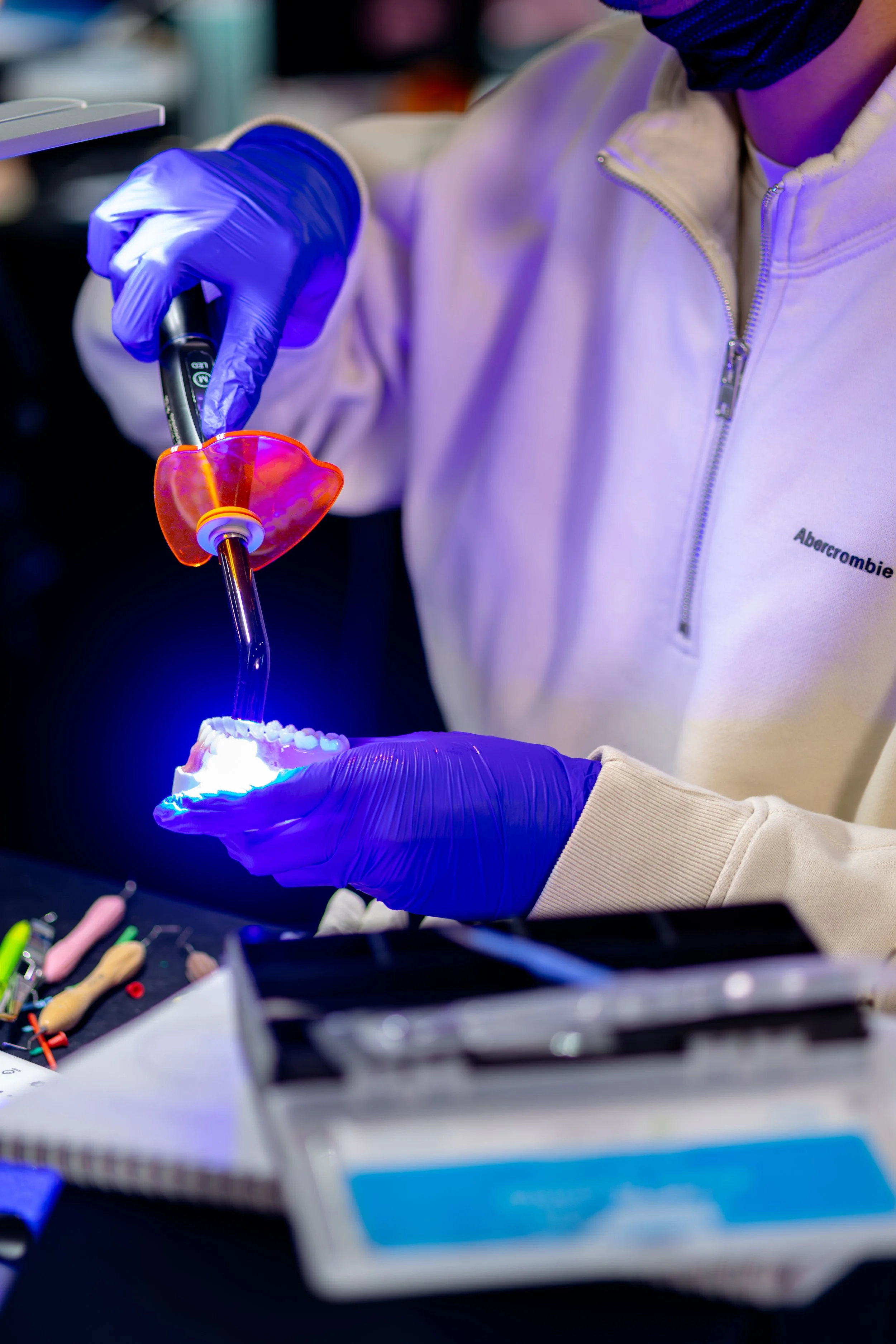 A person wearing purple gloves and a beige jacket with a zip working on a dental model under blue light.