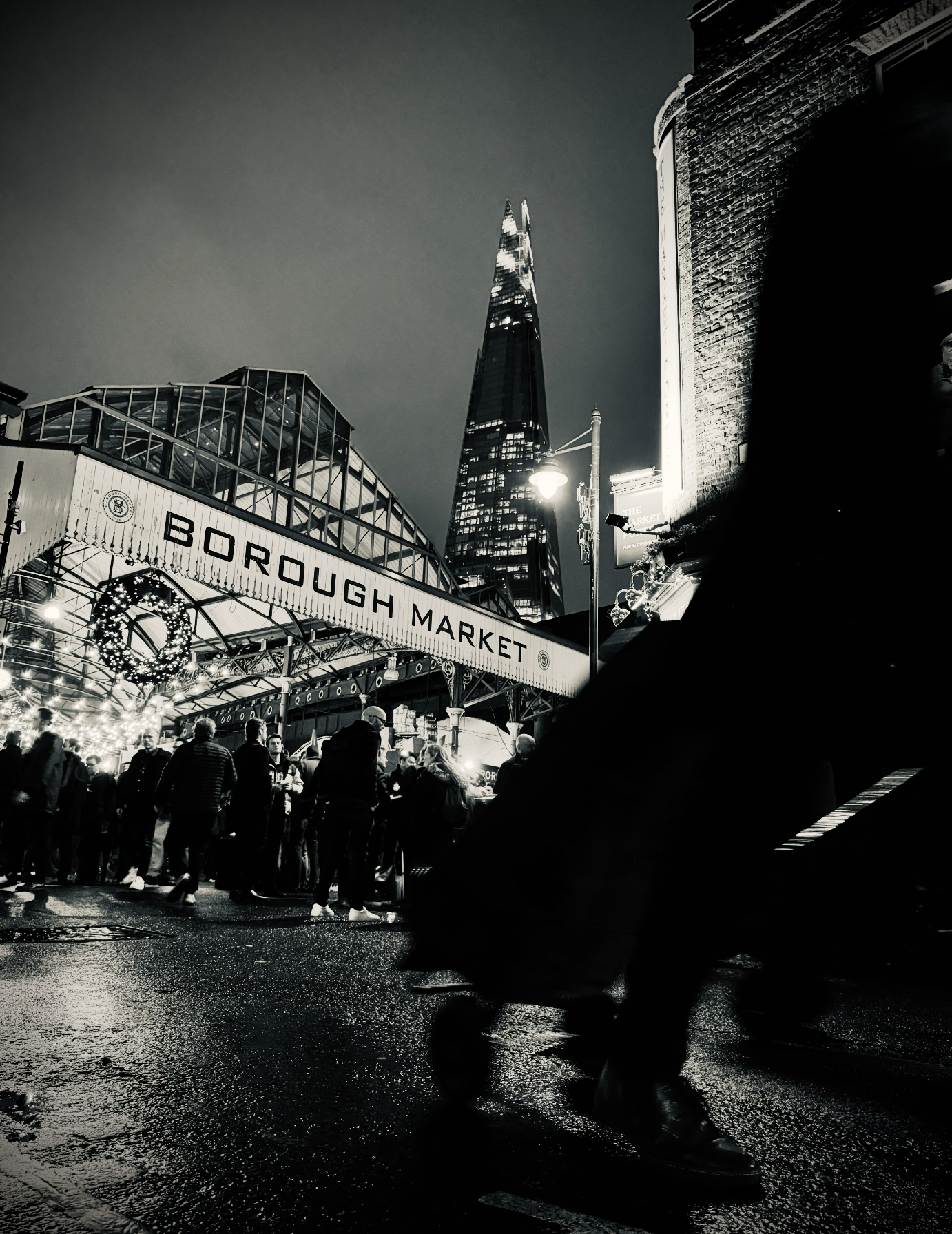 Night scene at Borough Market in London with people walking and the Shard skyscraper in the background, black and white photo.