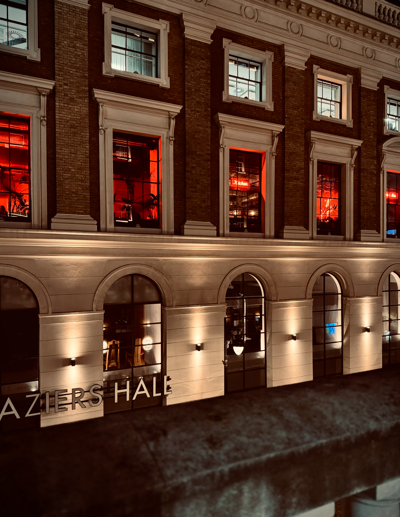 Nighttime view of a building with illuminated red and white interior lighting, brick exterior, large arched windows, and signs that read 'AZIERS HALL'.