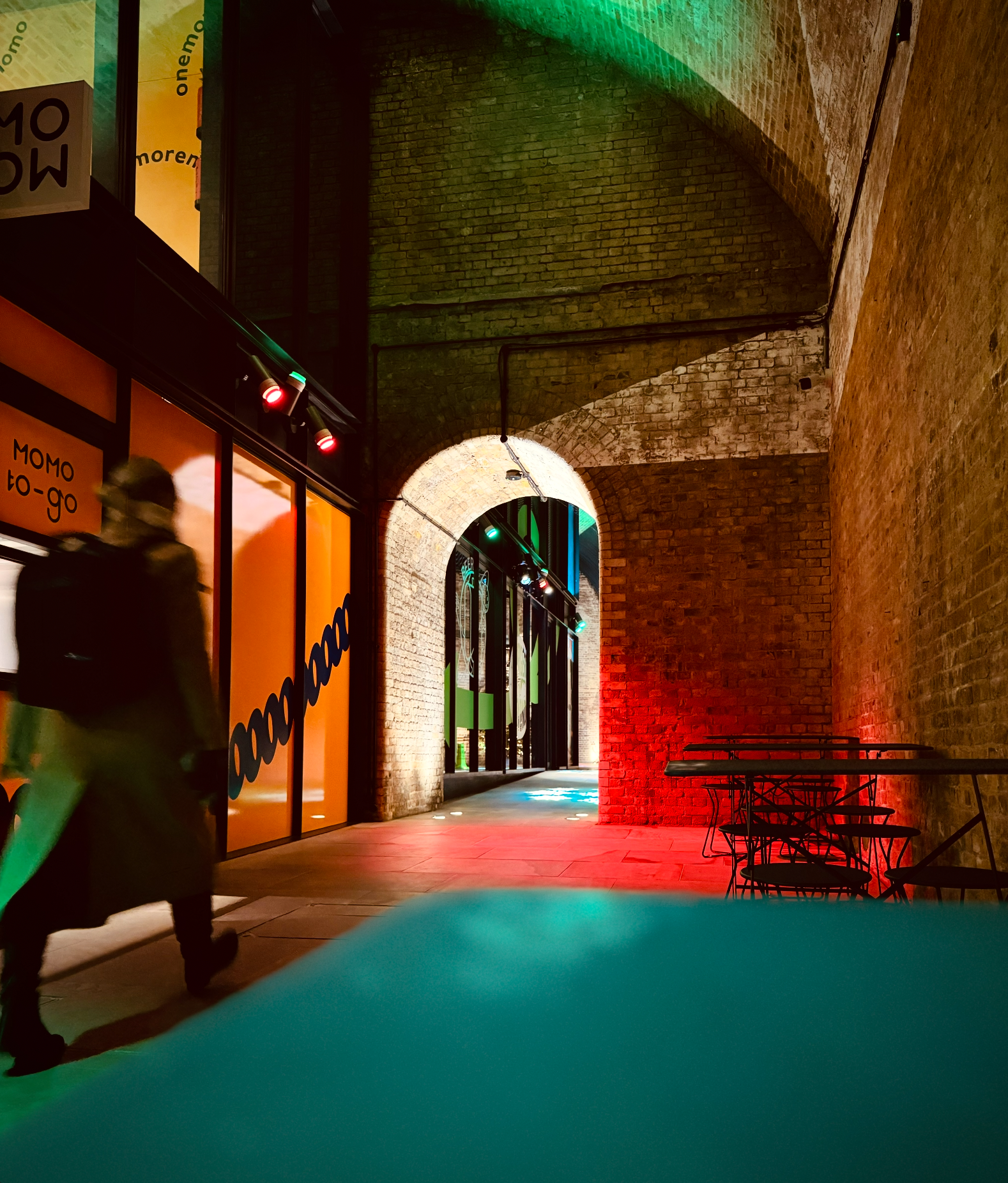 An underground passageway with brick walls and arches, illuminated with multicolored lights. On the left, there is a large orange window with black text reading 'MOMO to-go,' and a person is walking past. On the right, there are empty tables and chai