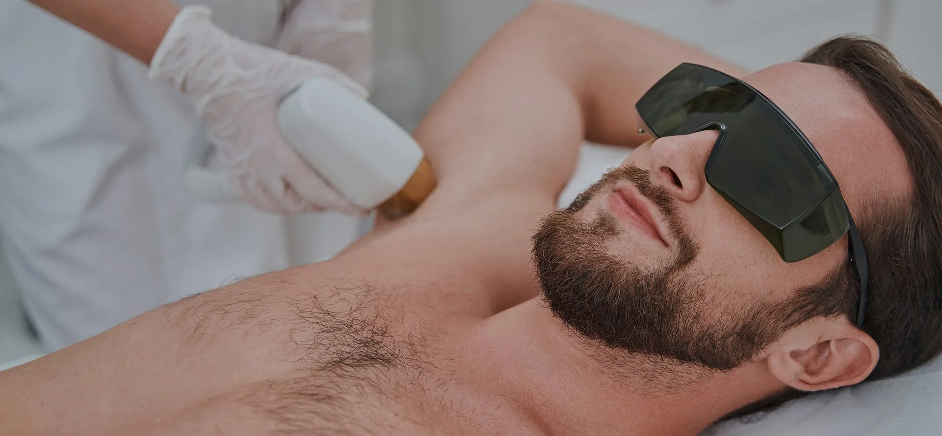 Illustration d'une séance d'épilation laser des aisselles chez un homme portant des lunettes de protection, avec une pièce à main appliquée sous le bras. Prestation réalisée chez Eminails à Waremme, près de Liège.