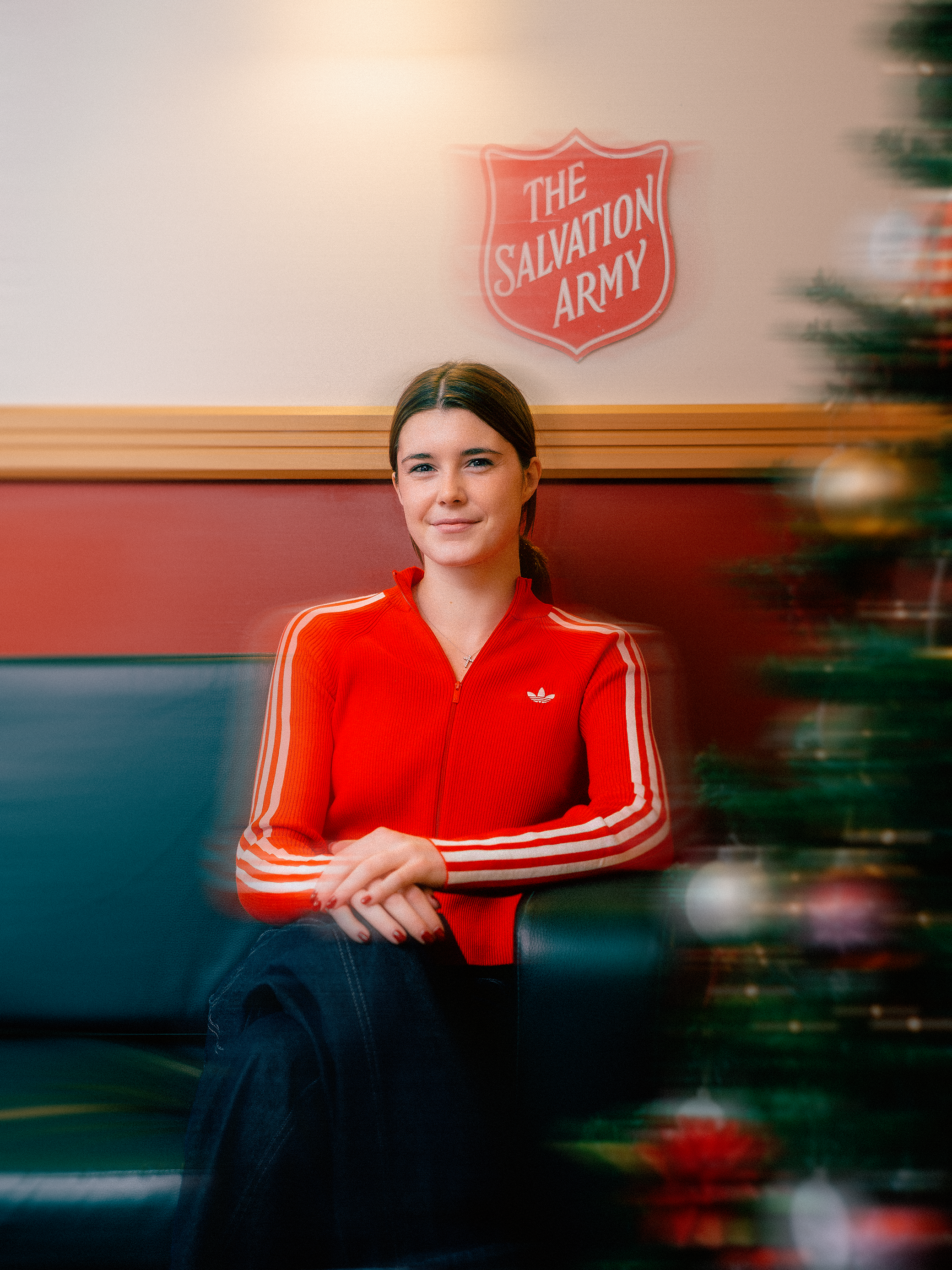 A young woman sitting on a green bench with a Christmas tree on her right. She is wearing a red hoodie with striped sleeves and a small logo. Behind her on the wall is a sign for The Salvation Army.