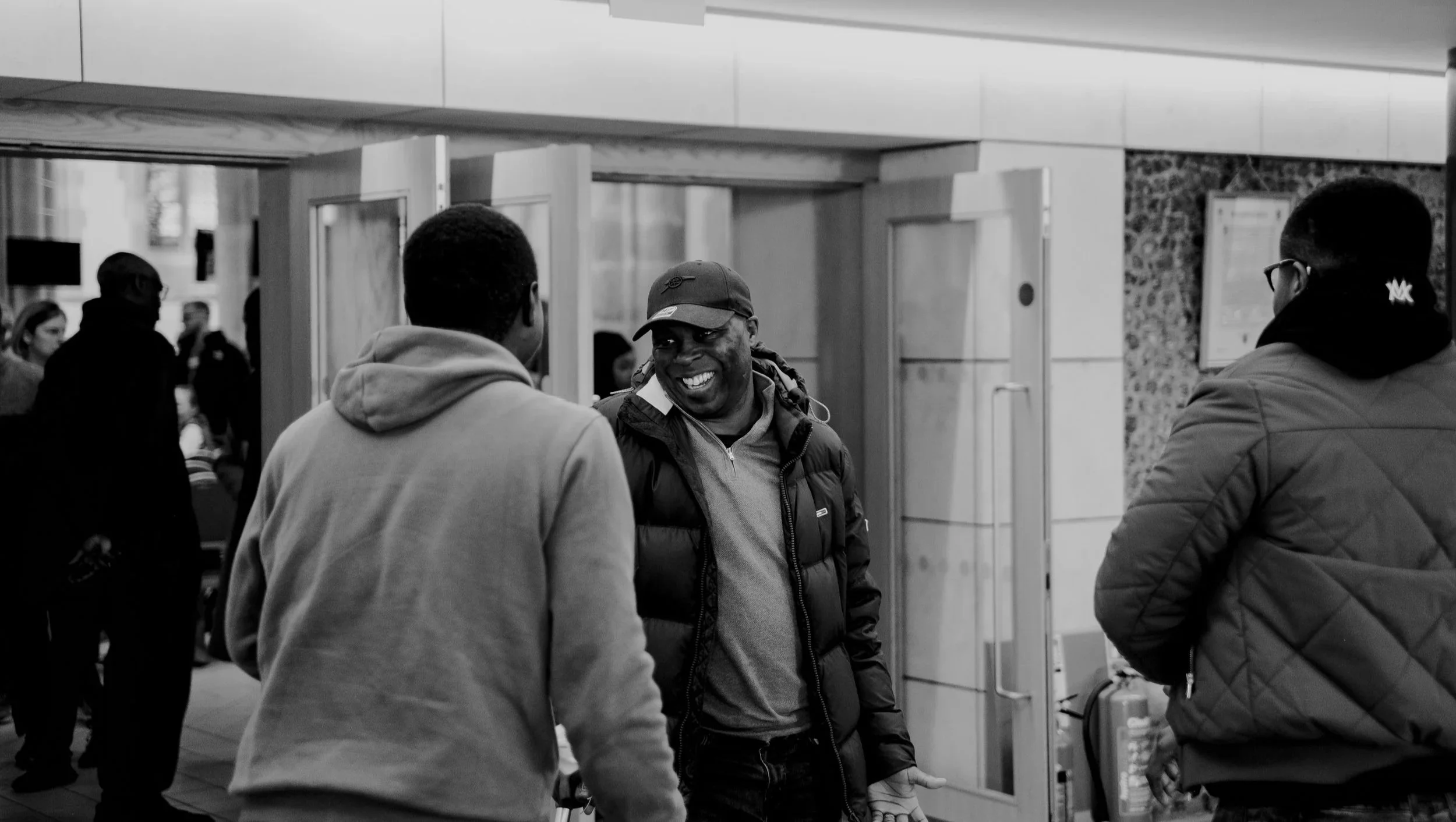 Three men talking and smiling at an airport terminal, with other travelers in the background