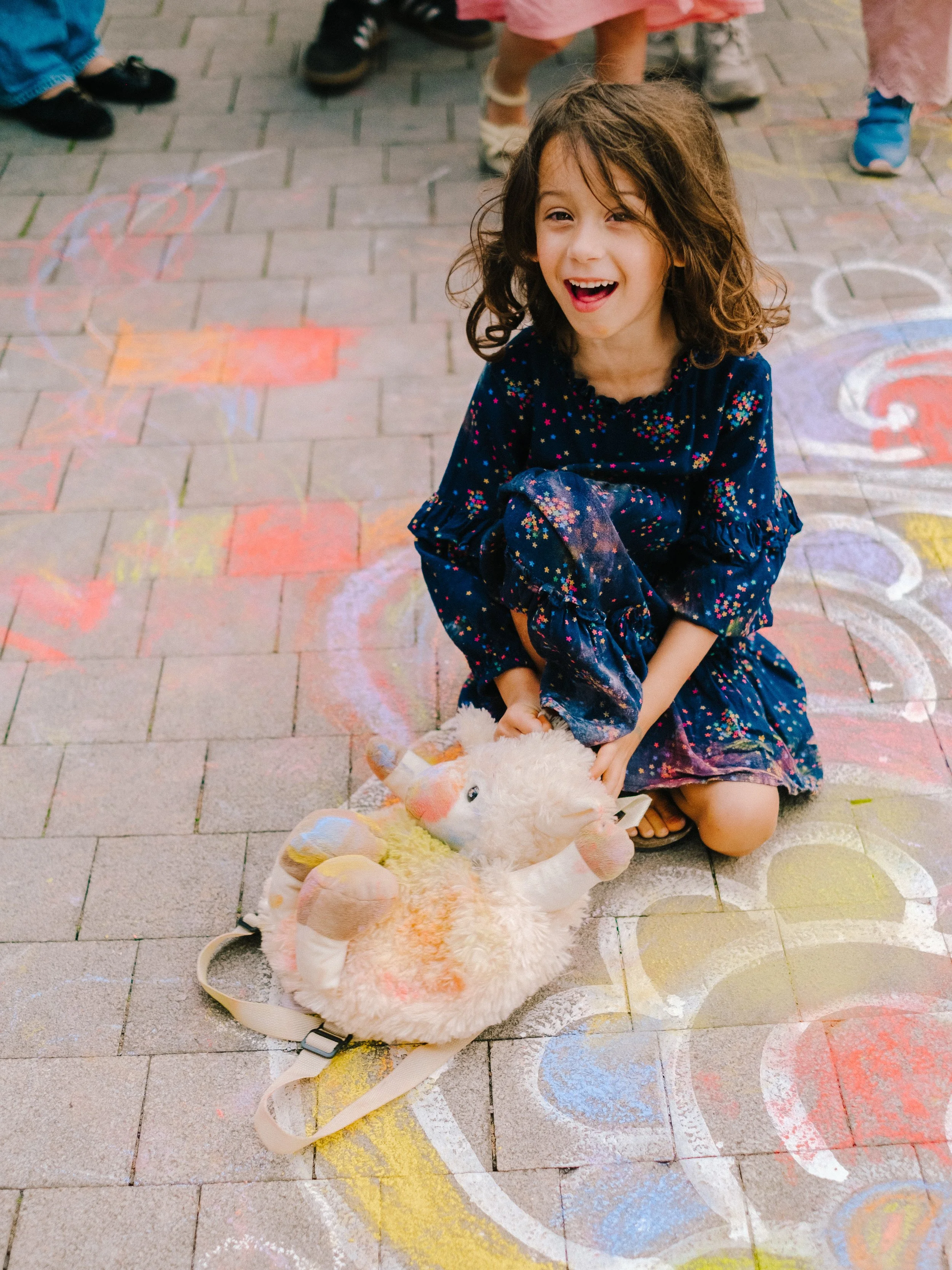A young girl with curly brown hair wearing a navy blue dress with colorful floral patterns, smiling and sitting on a brick pavement surrounded by colorful chalk drawings. She is holding a plush unicorn toy.