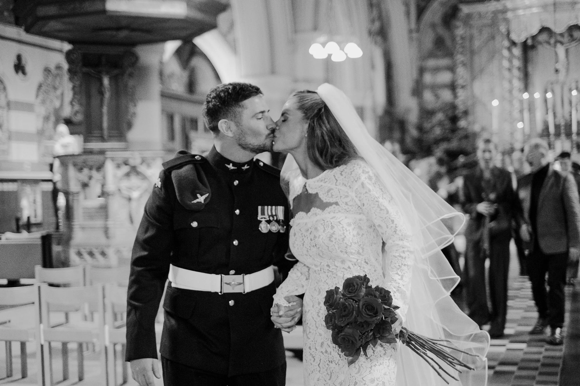 A wedding ceremony showing a bride and groom kissing in a church, with guests watching in the background. The groom is in the military uniform, and the bride is wearing a lace wedding dress holding a bouquet of roses.