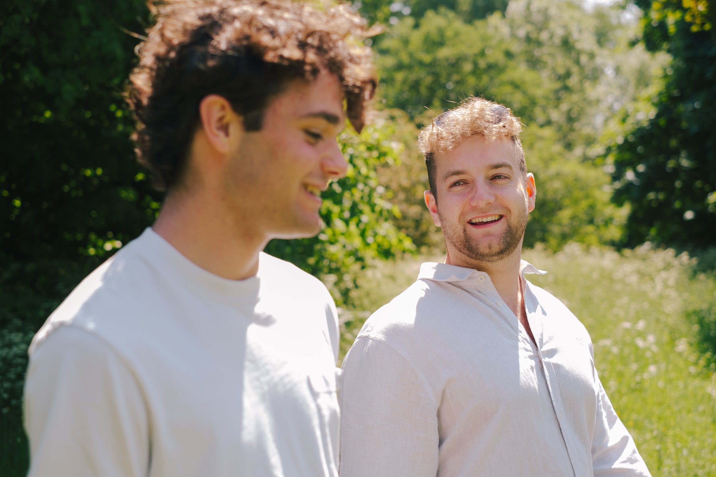 Two young men are outdoors on a sunny day, walking and talking with trees in the background.