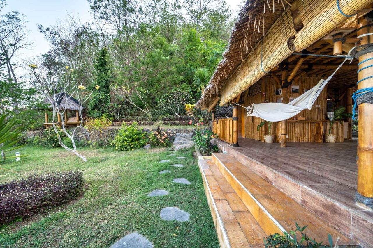 Tropical bamboo house with a hammock on the porch, surrounded by lush greenery and trees, with a stone path leading to the entrance.