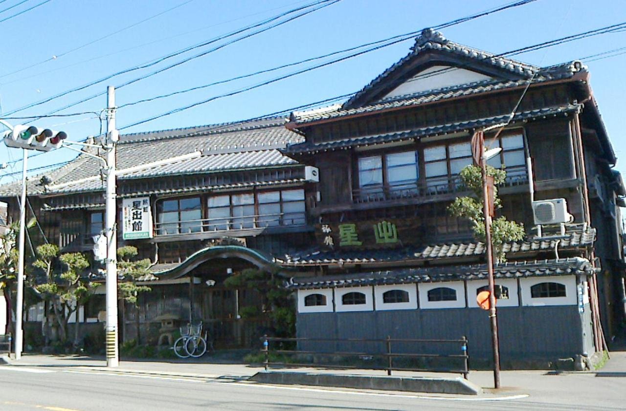 Traditional Japanese building with tiled roof, wooden facade, and signage in Japanese characters. Located on a street with traffic signals and power lines.