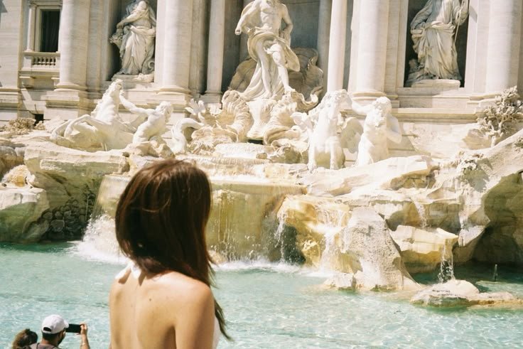 Woman looking at Trevi Fountain in Rome, Italy, with sculptures and cascading water, and a person taking a photo.