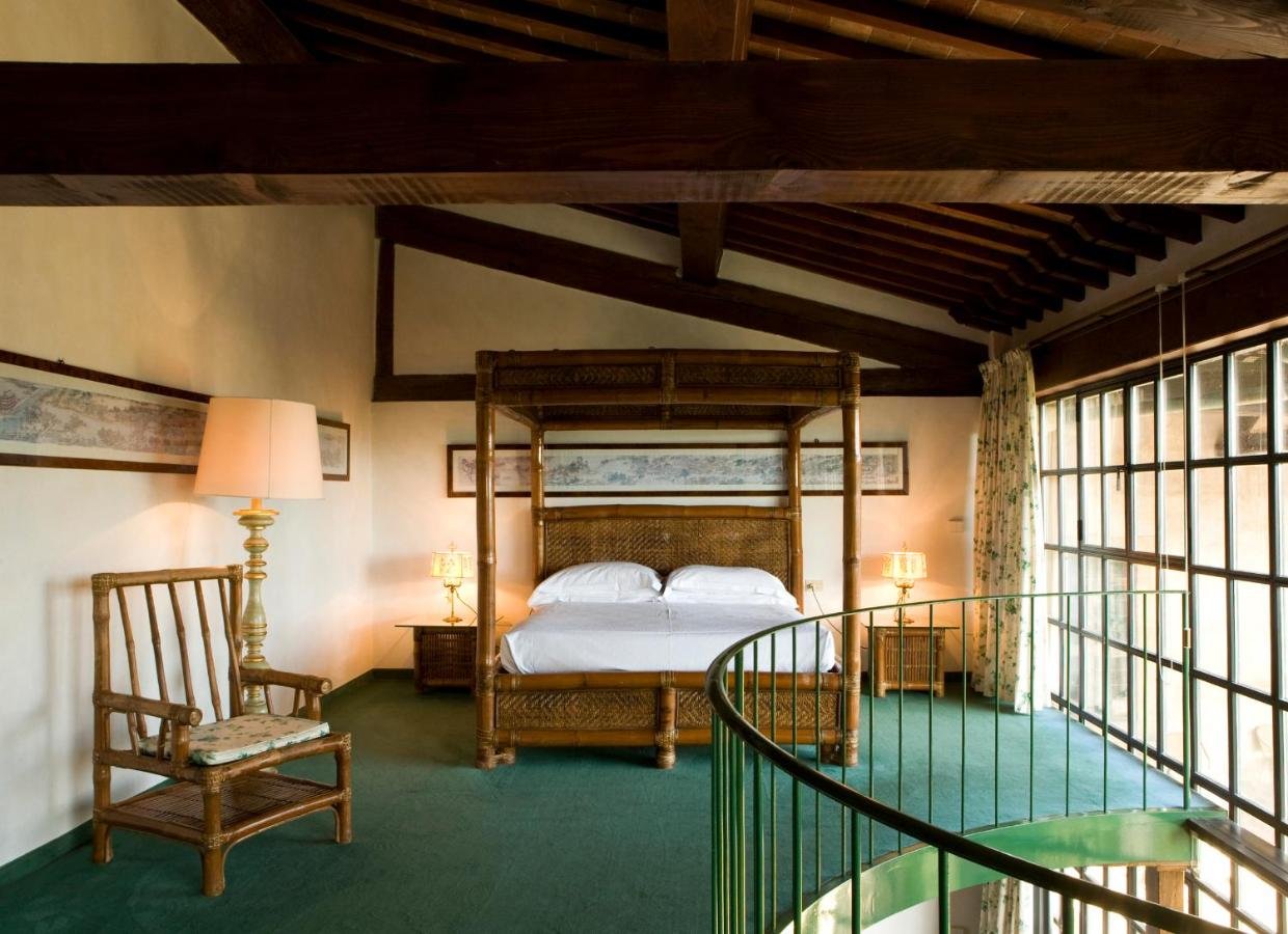 Elegant bedroom with a canopy bed, wooden beams, and green carpet. Features a wooden chair, bedside tables, lamps, framed art on the walls, and large windows with floral curtains, overlooking a spiral staircase.