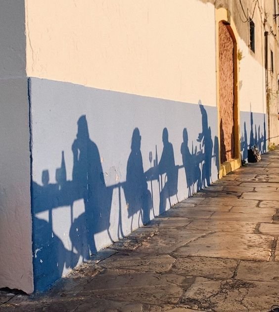 Shadows of people dining on a wall at sunset, showing silhouettes of tables, chairs, and individuals on an outdoor patio.
