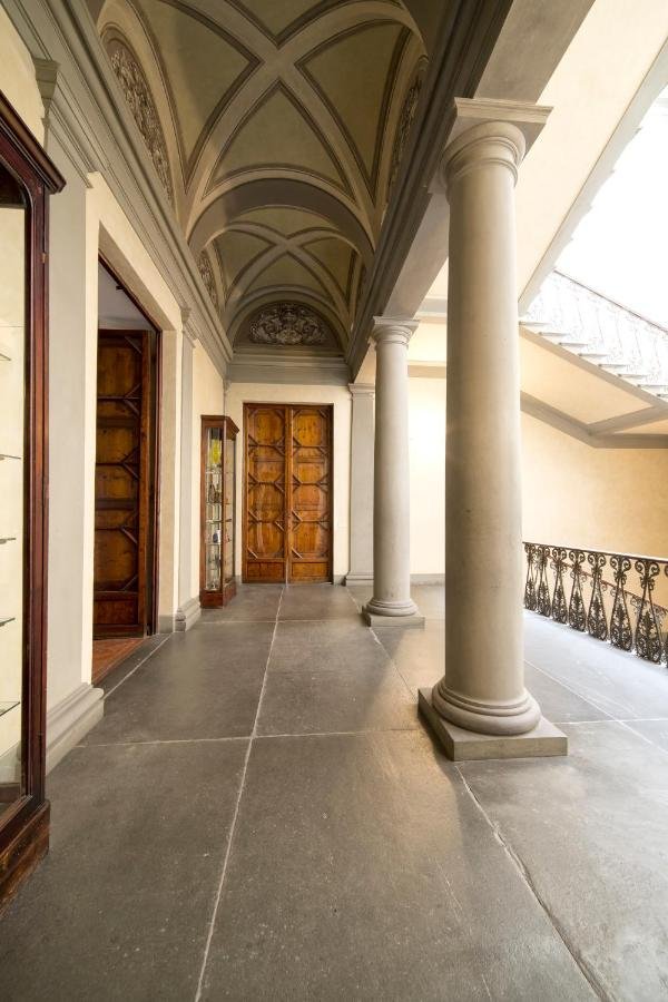 Interior of a historical building with arched ceiling and stone columns. The floor is tiled, and wooden doors lead to separate rooms. A metal railing lines the staircase, visible on the right.