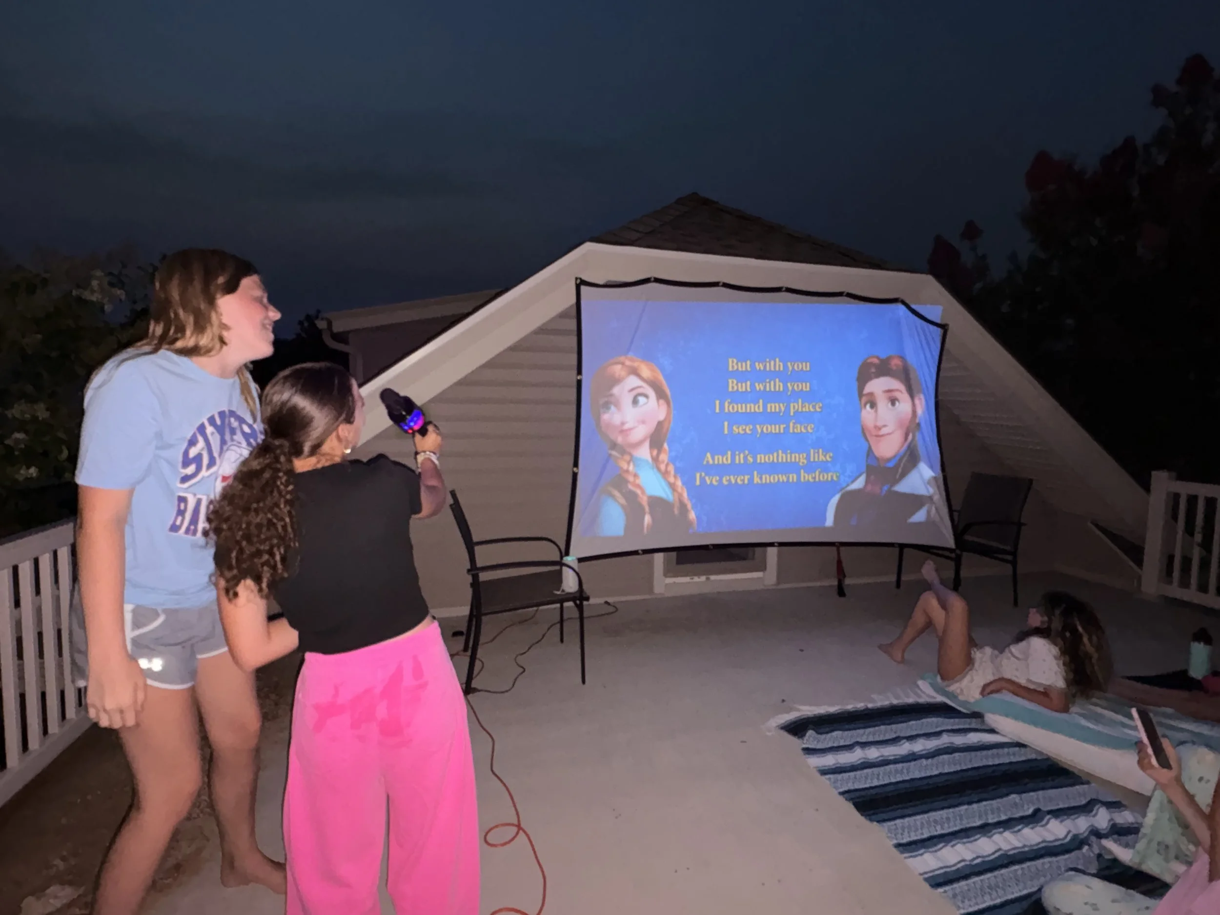 People gathered outside watching a projection of a scene from the movie 'Frozen' on a screen, with lyrics displayed. A girl is lying on a blanket on the ground, and other children and teens are standing nearby, some with a microphone.