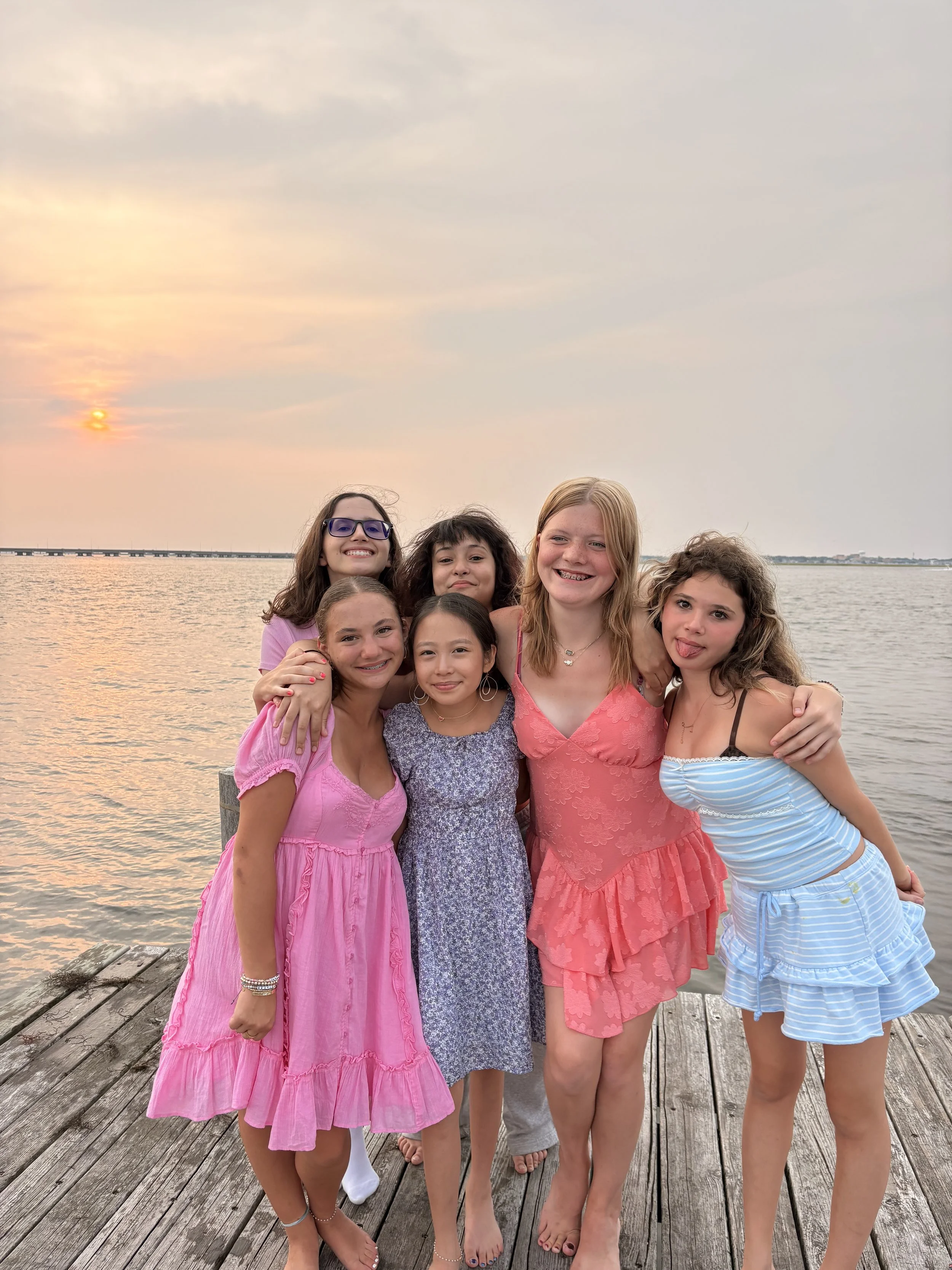 A group of seven young girls standing close together on a wooden dock by the water during sunset, smiling and posing for a photo.