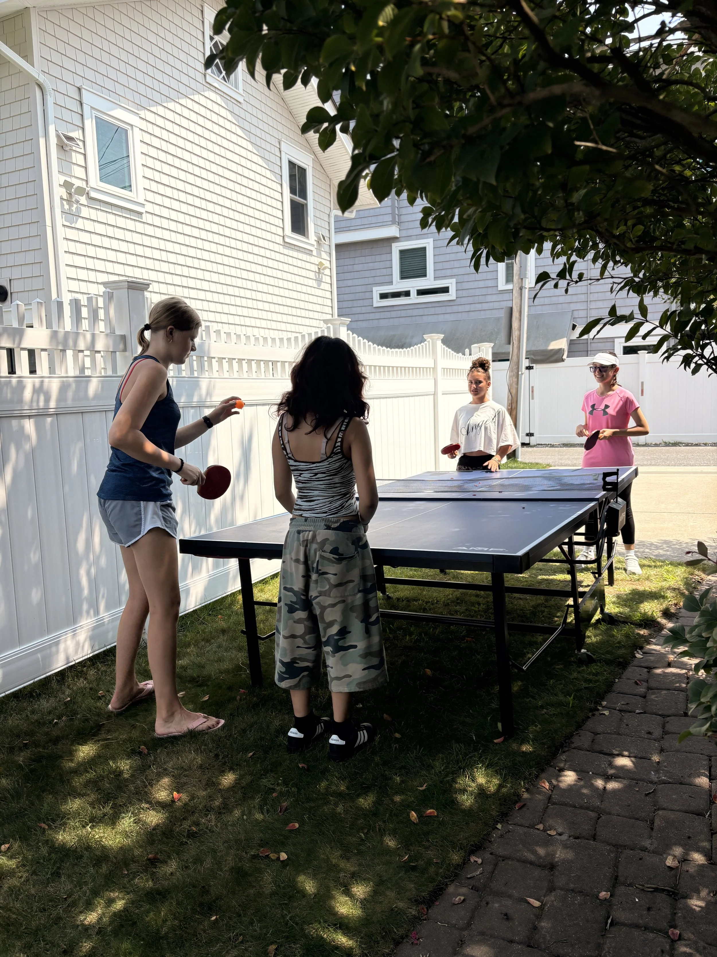 Four young women are playing ping pong outdoors in a backyard, with a white house and a white fence nearby. One woman is about to serve, while the others are watching and waiting. The scene is shaded by trees.
