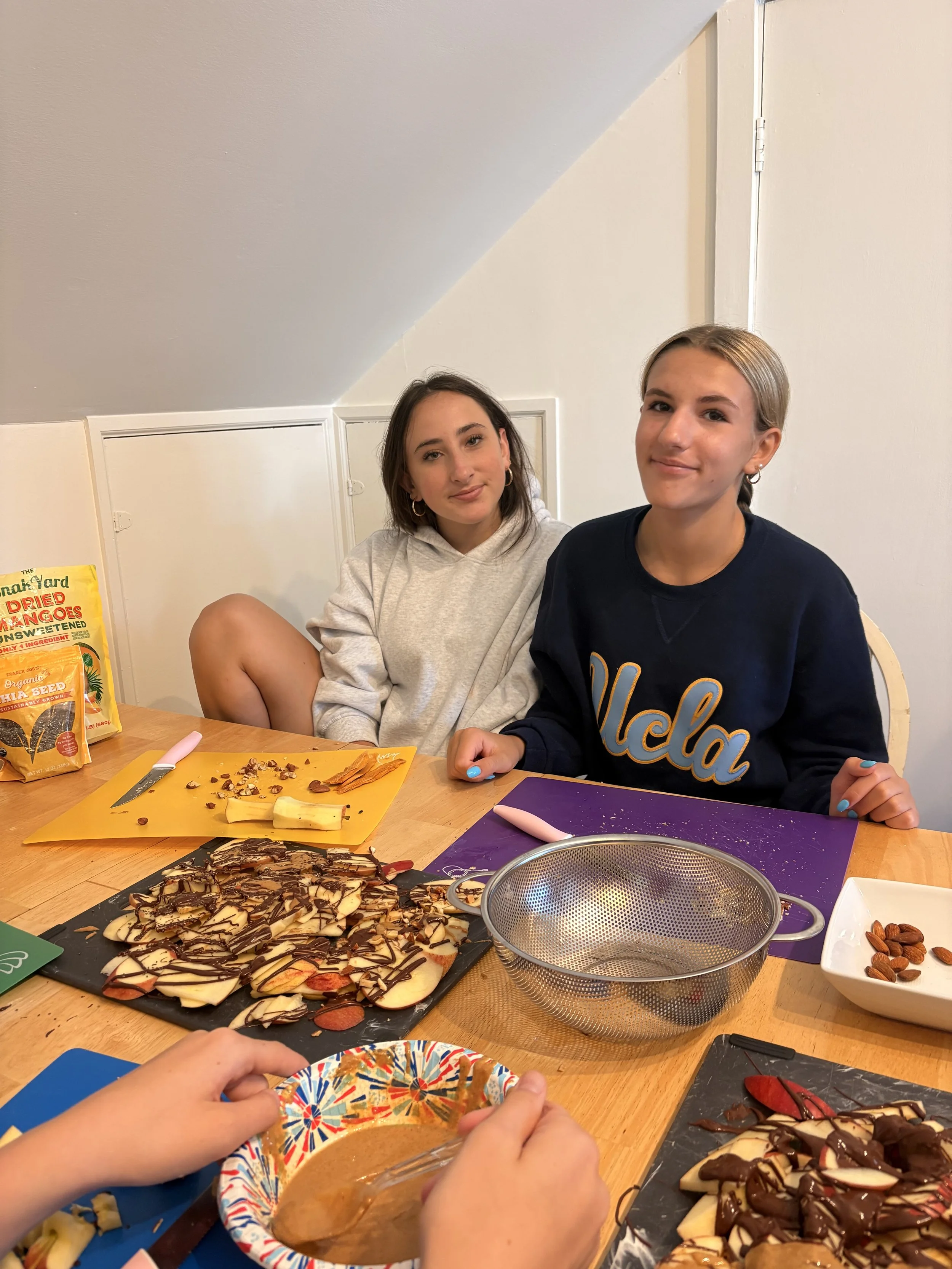 Two young women sitting at a table filled with food, including a large chocolate-drizzled pizza, bowls of nuts, and chocolate cake, in a casual indoor setting. One woman is wearing a white hoodie and the other a dark sweatshirt with 'UCLA' on it.