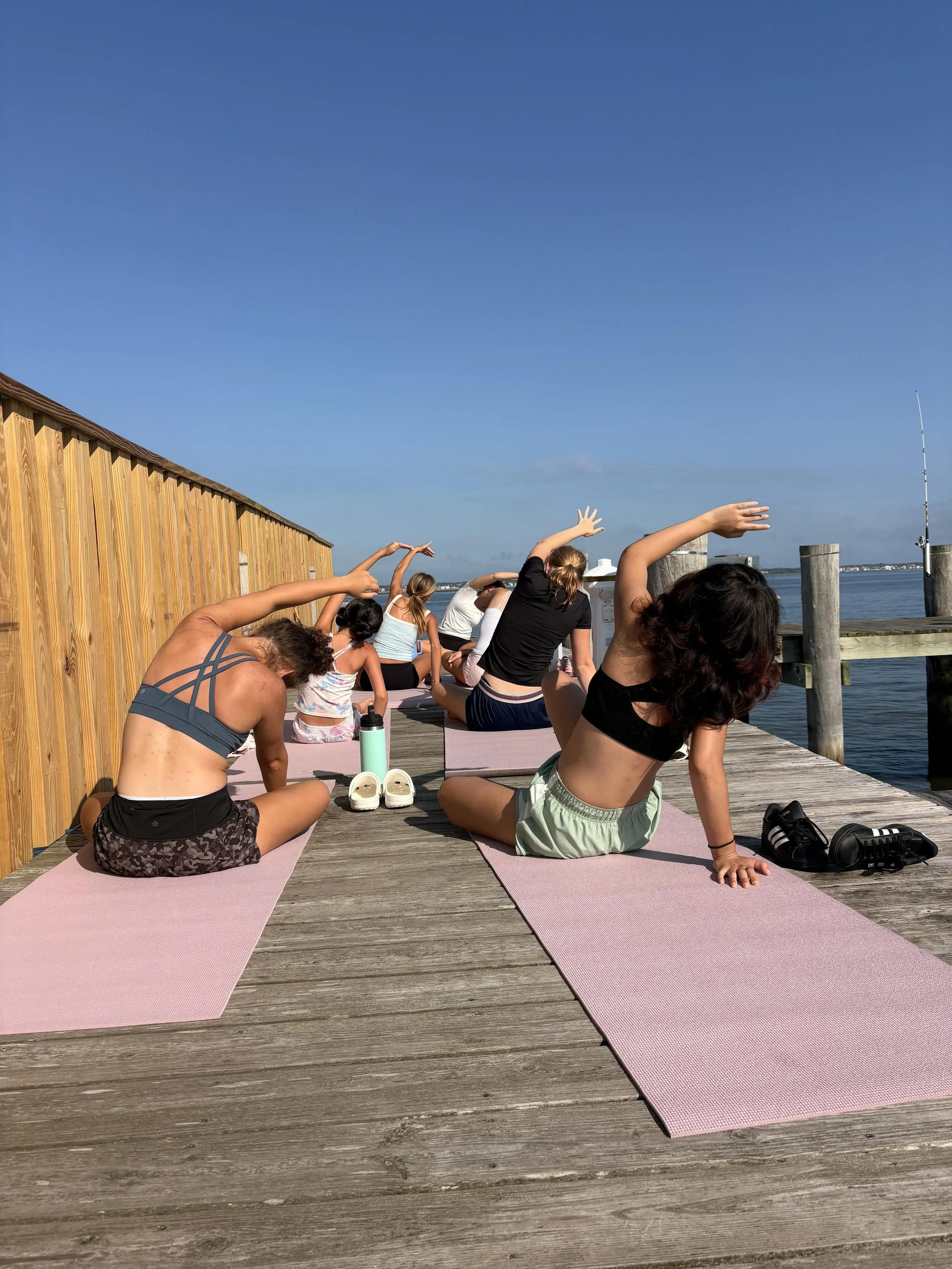 People practicing yoga on a wooden dock by the water, with a clear blue sky and fishing rod in the background.