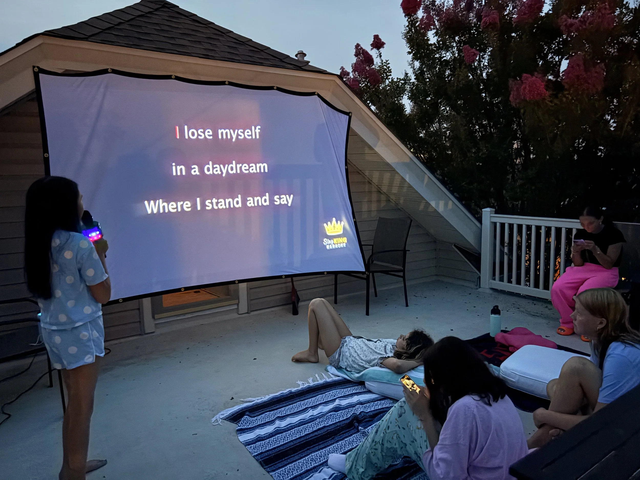 People, mostly children, sitting and lying on the ground outdoors at dusk, watching a screen displaying karaoke lyrics: 'I lose myself in a daydream where I stand and say.' Some children are holding phones or glow sticks.