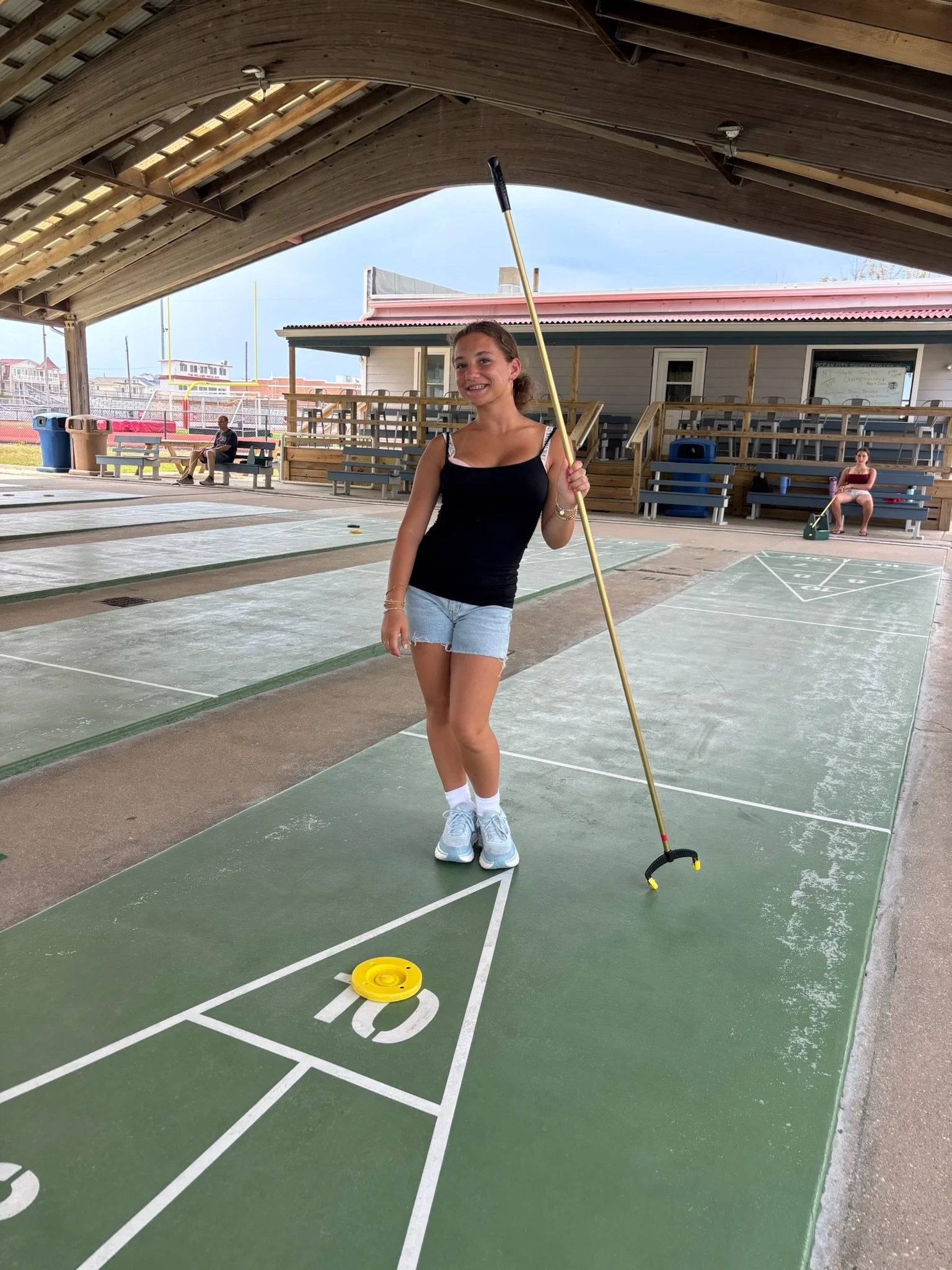 Young woman playing shuffleboard at an outdoor courts with other people in the background.