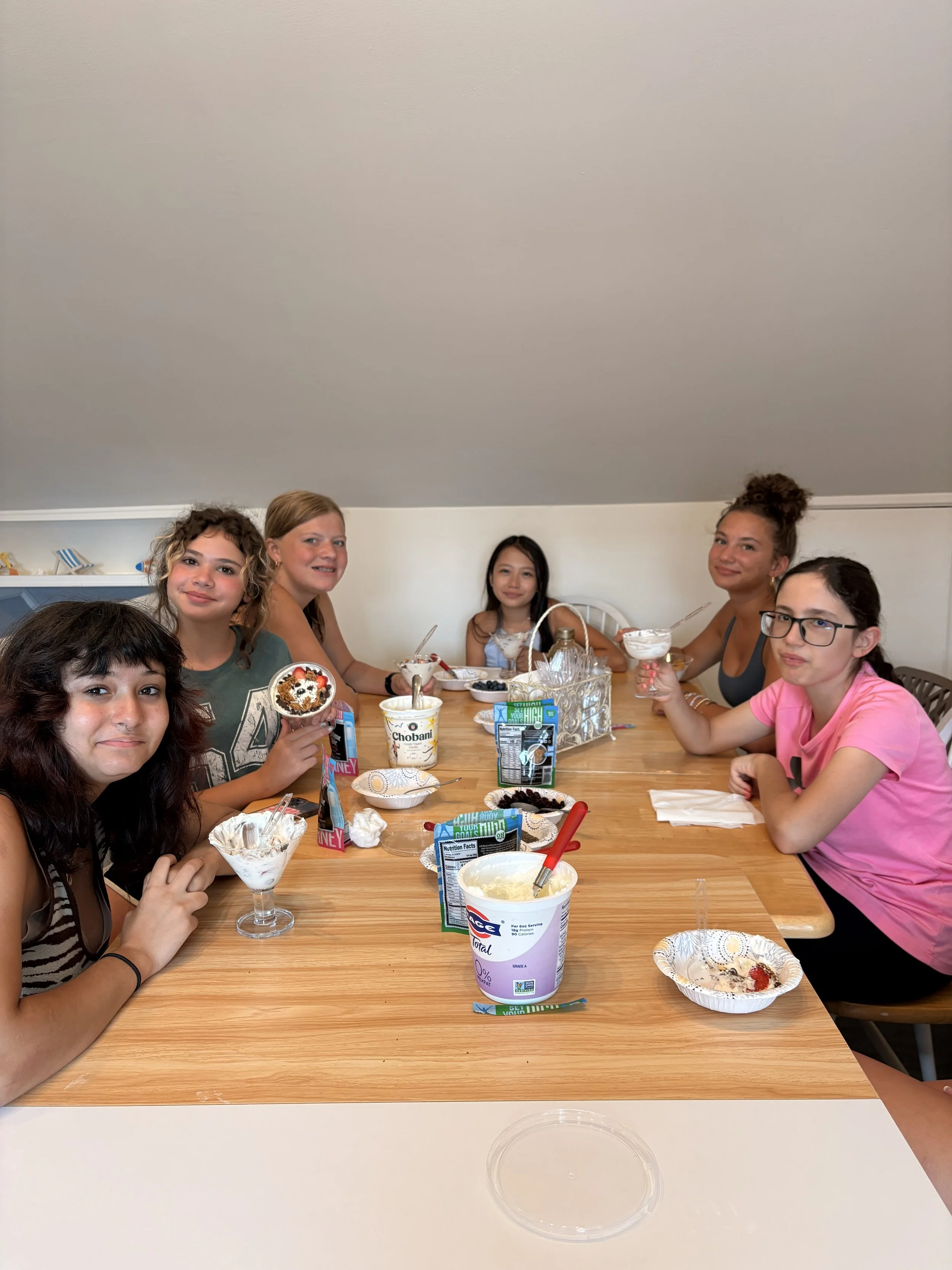 Six people sitting at a wooden dining table enjoying ice cream dessert with various toppings, surrounded by bowls and snack packages.
