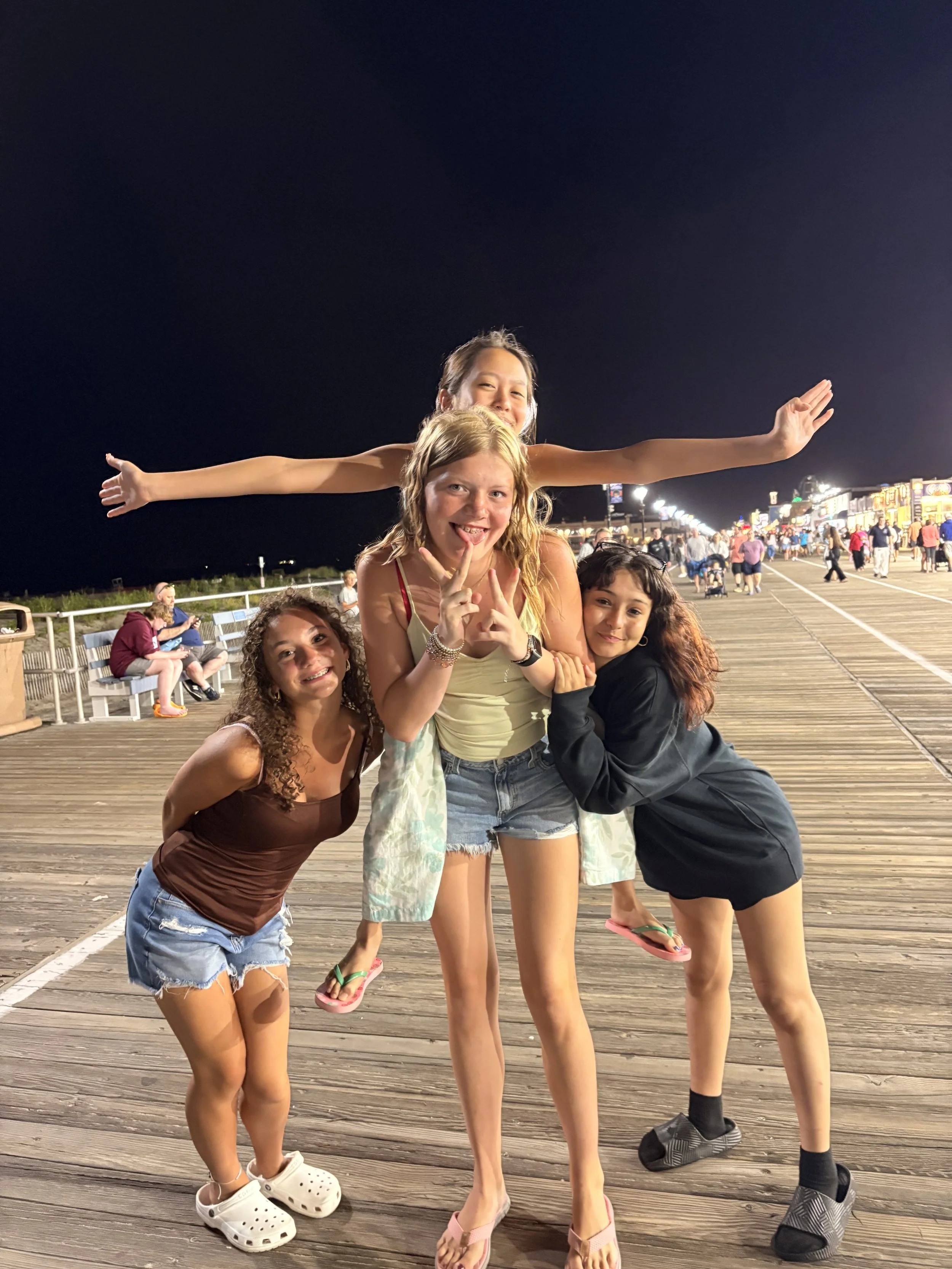 Group of five girls smiling and posing on a wooden pier at night, with amusement park lights in the background. One girl is lying across the shoulders of two others, extending her arms out.
