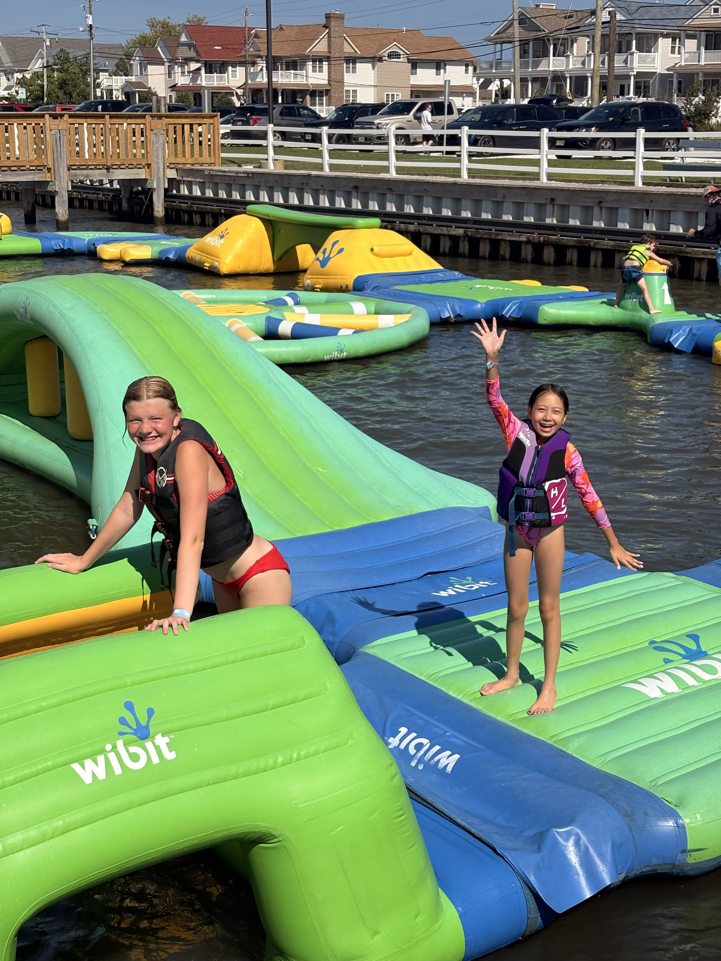 Children playing and jumping off an inflatable water obstacle course in a body of water, with adults supervising nearby. Houses and parked cars are in the background.